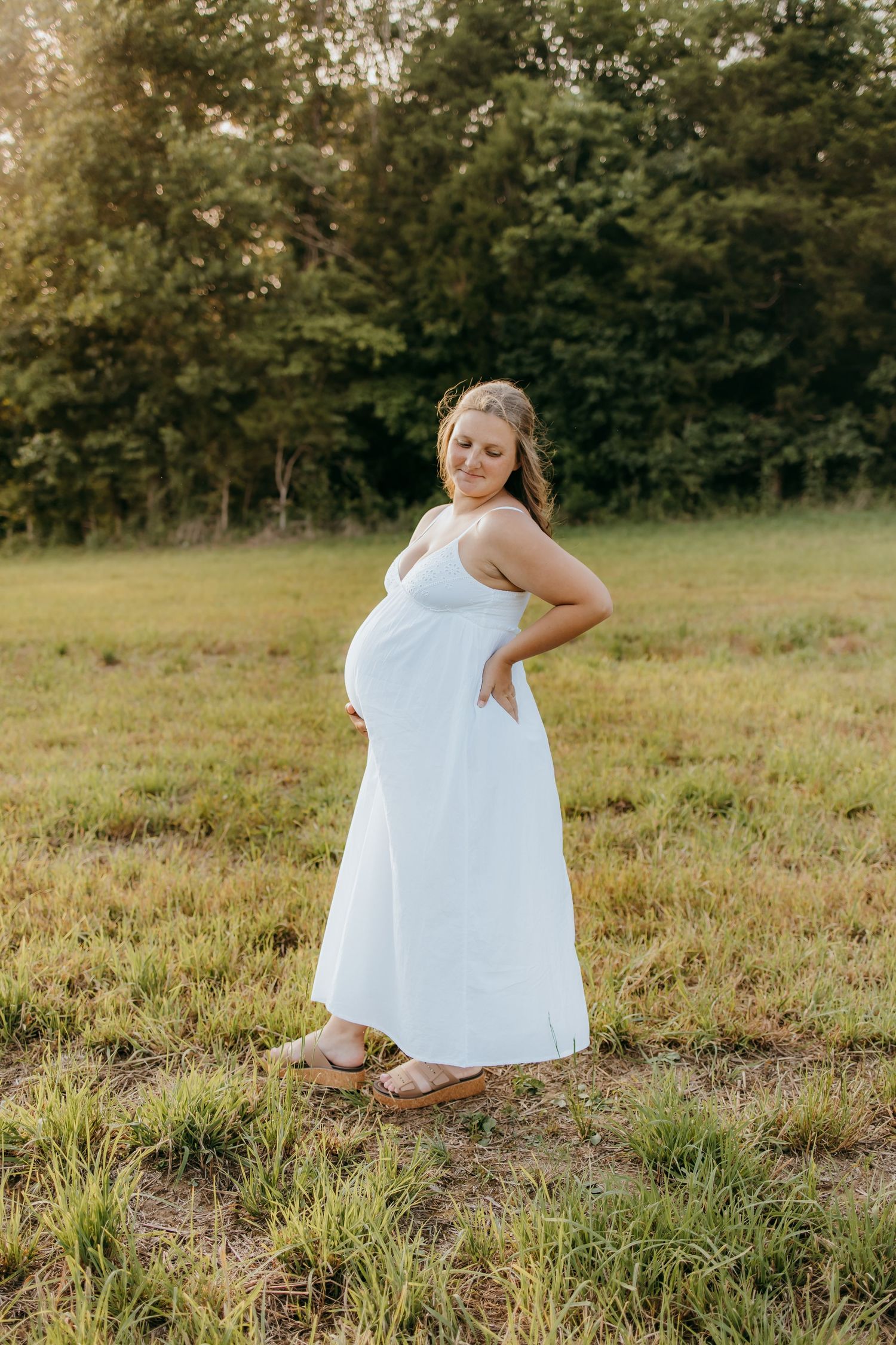 Pregnant woman in white maxi dress posing in a sunlit field with trees in background during golden hour.