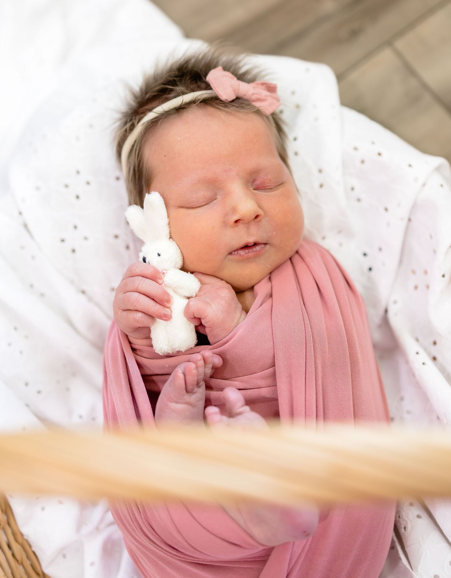 Newborn baby girl swaddled in pink wrap holding a white bunny toy, wearing a pink bow headband, sleeping peacefully.