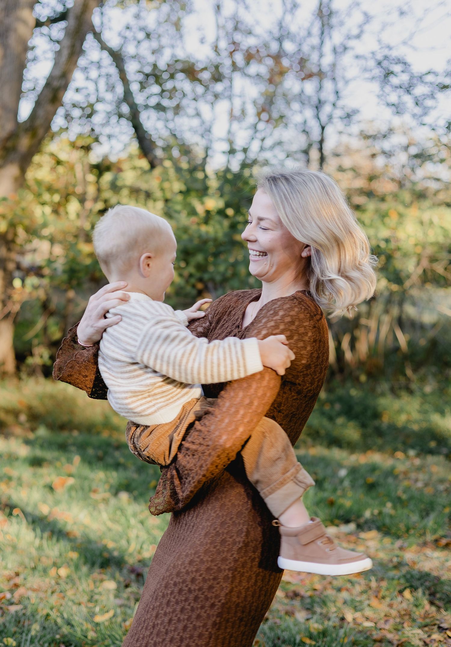 Smiling blonde woman holding a happy toddler outdoors in a sunny autumn garden, wearing a brown dress.