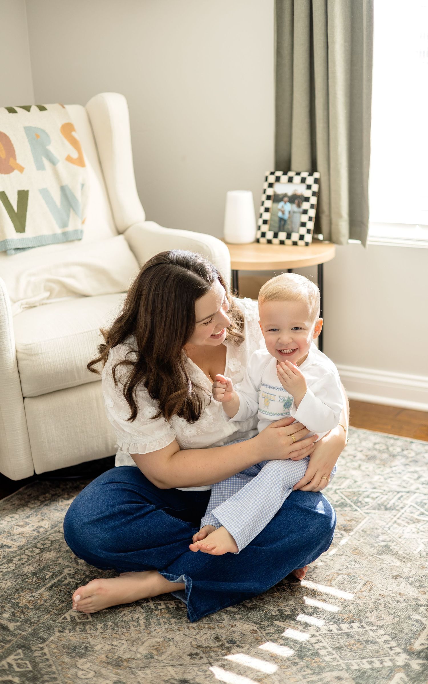 A mother sits cross-legged on a rug, smiling at her laughing toddler son in a bright, cozy nursery room.