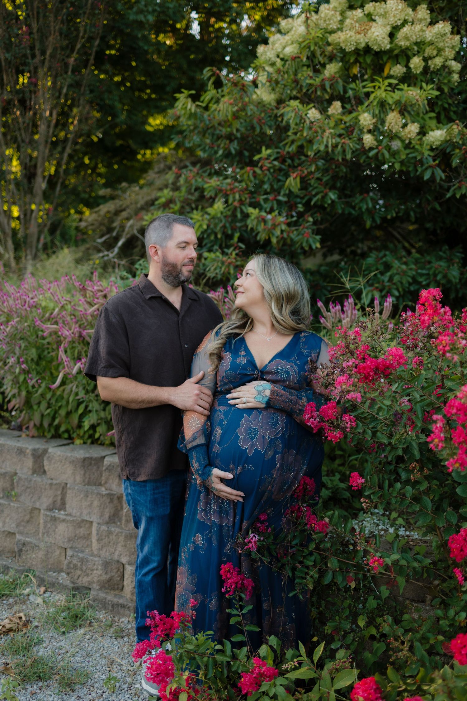 A couple shares a tender moment among vibrant pink flowers, the woman in a blue floral maternity gown cradling her baby bump.