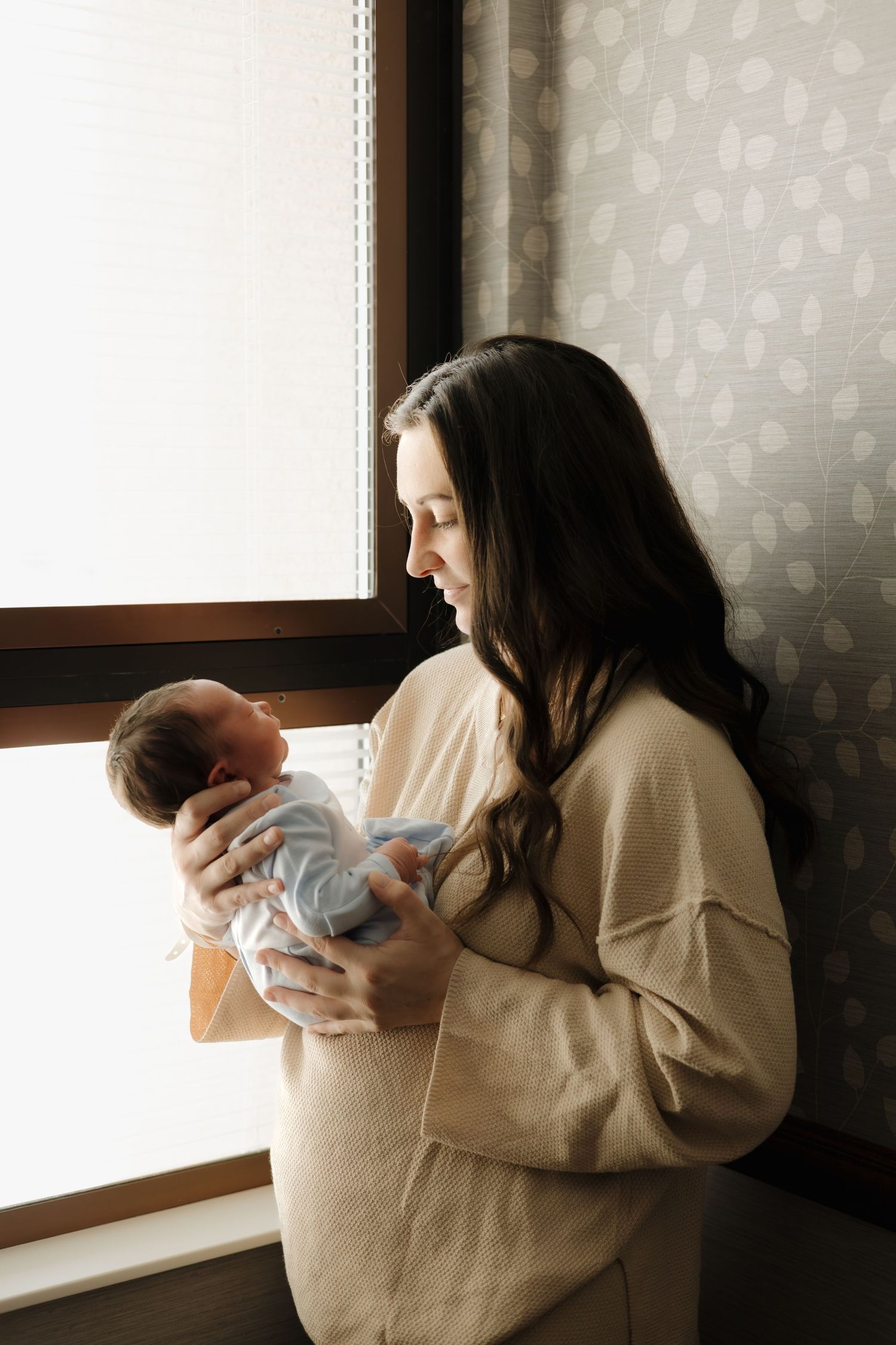 A smiling mother with long dark hair holds her newborn baby near a bright window in a cozy beige sweater.