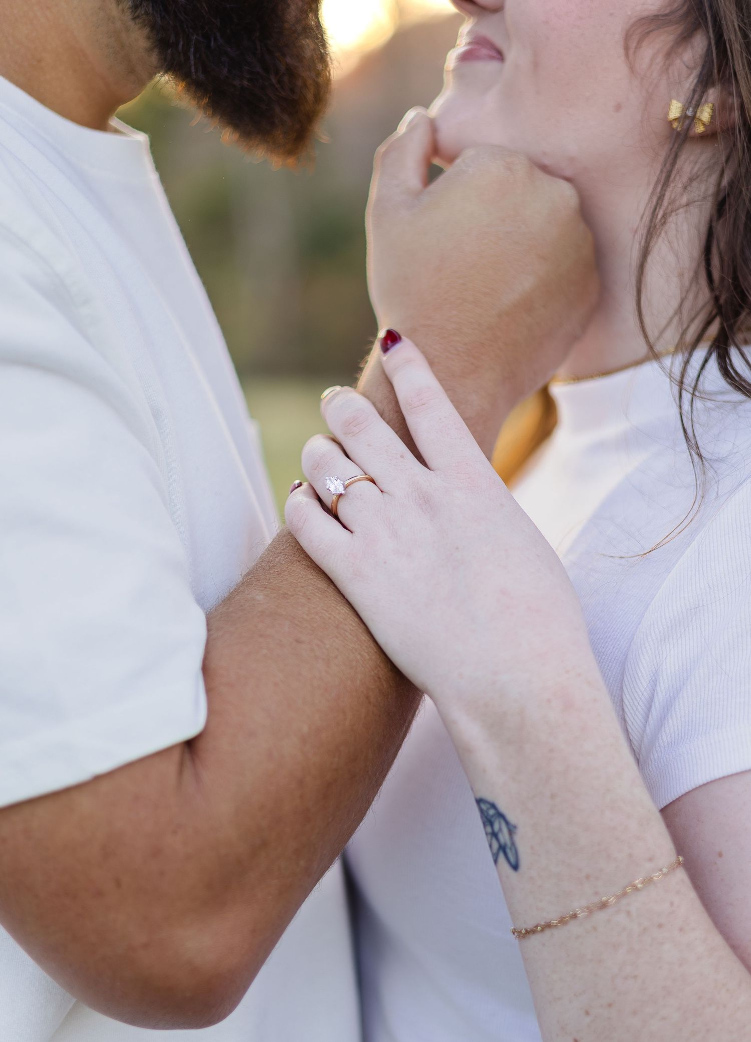 Close-up of a couple in white shirts, woman displaying an engagement ring while tenderly touching her partner's face.