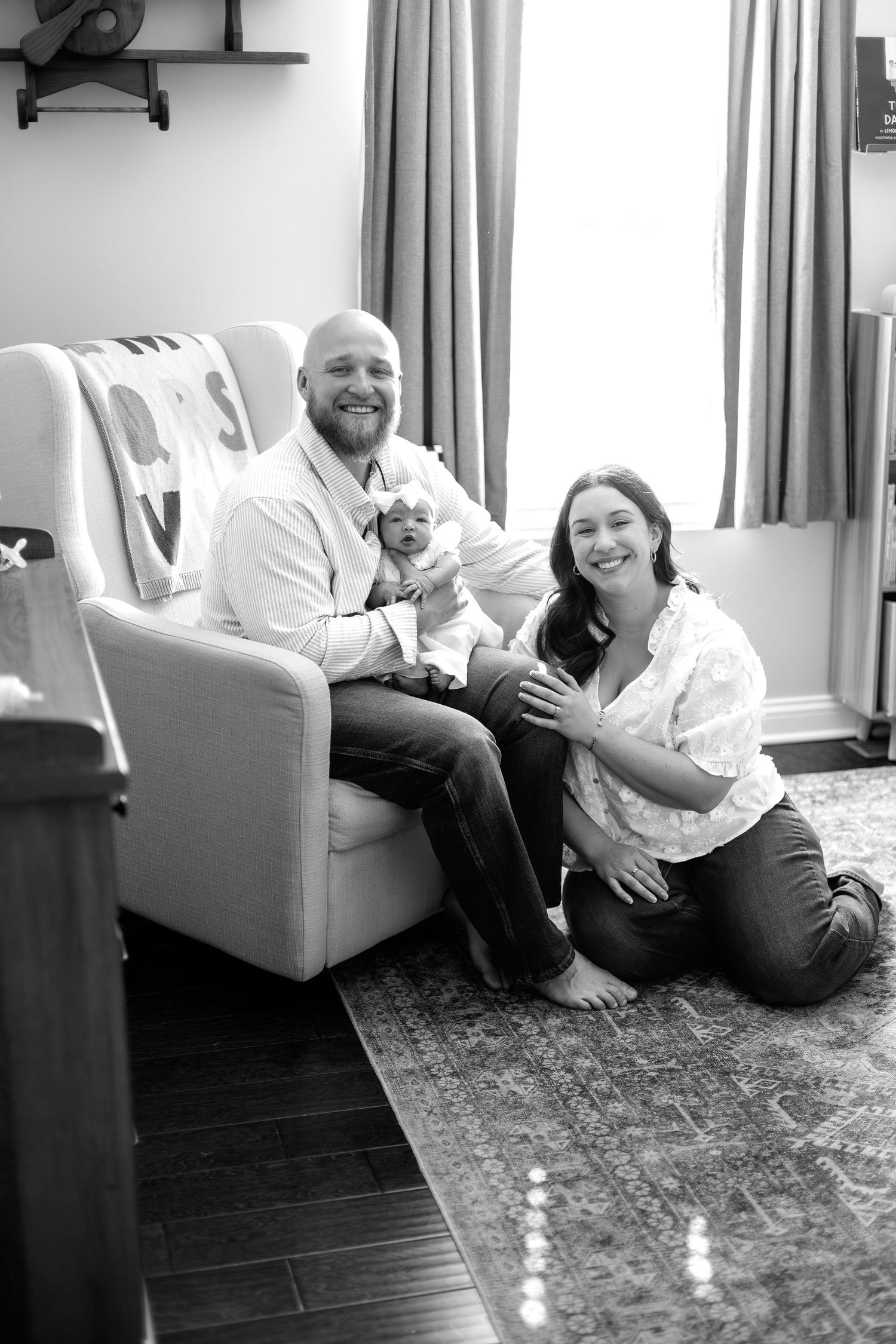 Black and white photo of smiling parents sitting with newborn baby in cozy nursery chair on patterned rug.