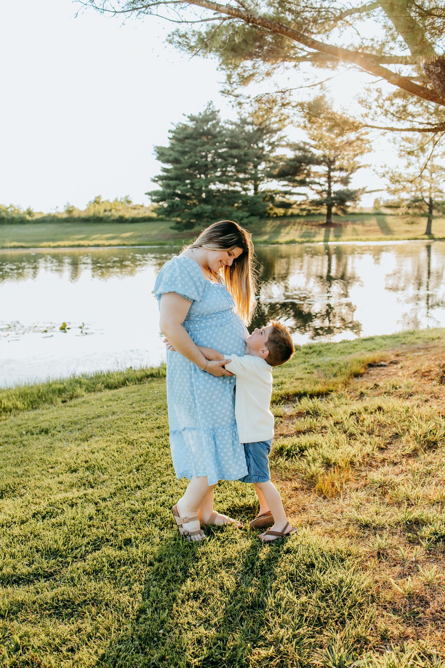 Pregnant woman in blue dress with young child hugging her belly near a serene lake at golden hour.
