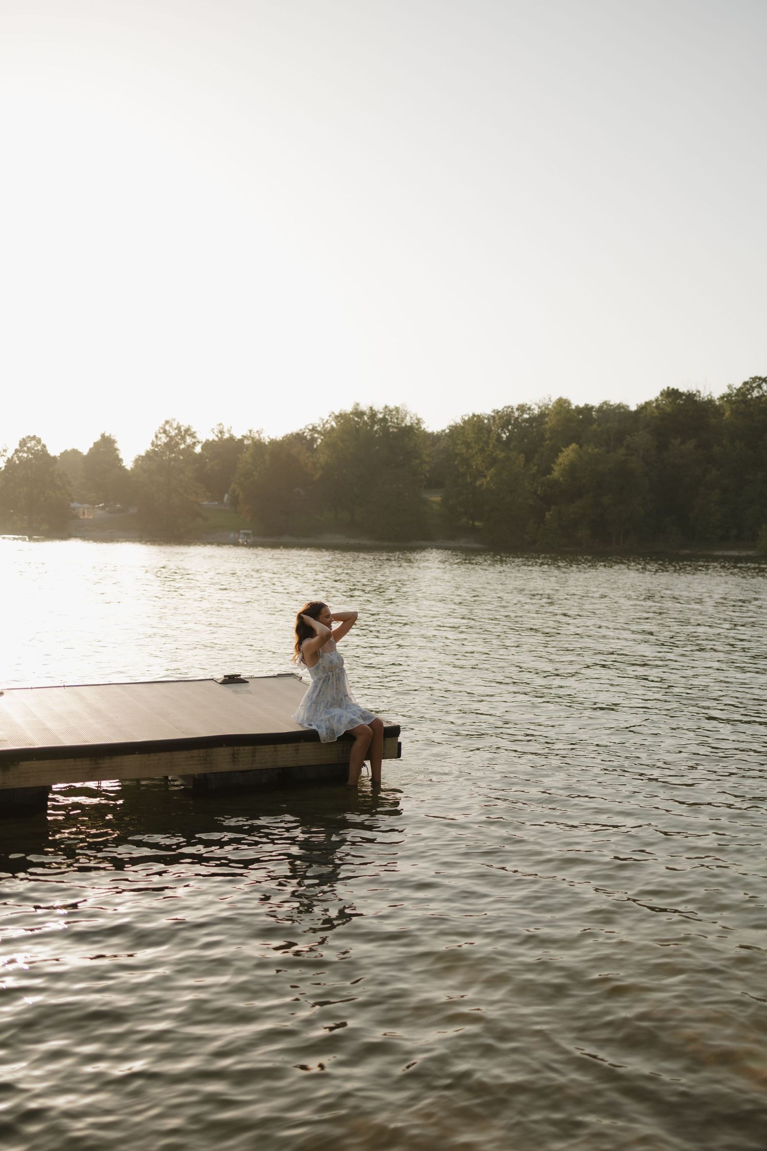 Senior in white dress sitting on wooden dock by calm lake at golden sunset with lush green trees in background.