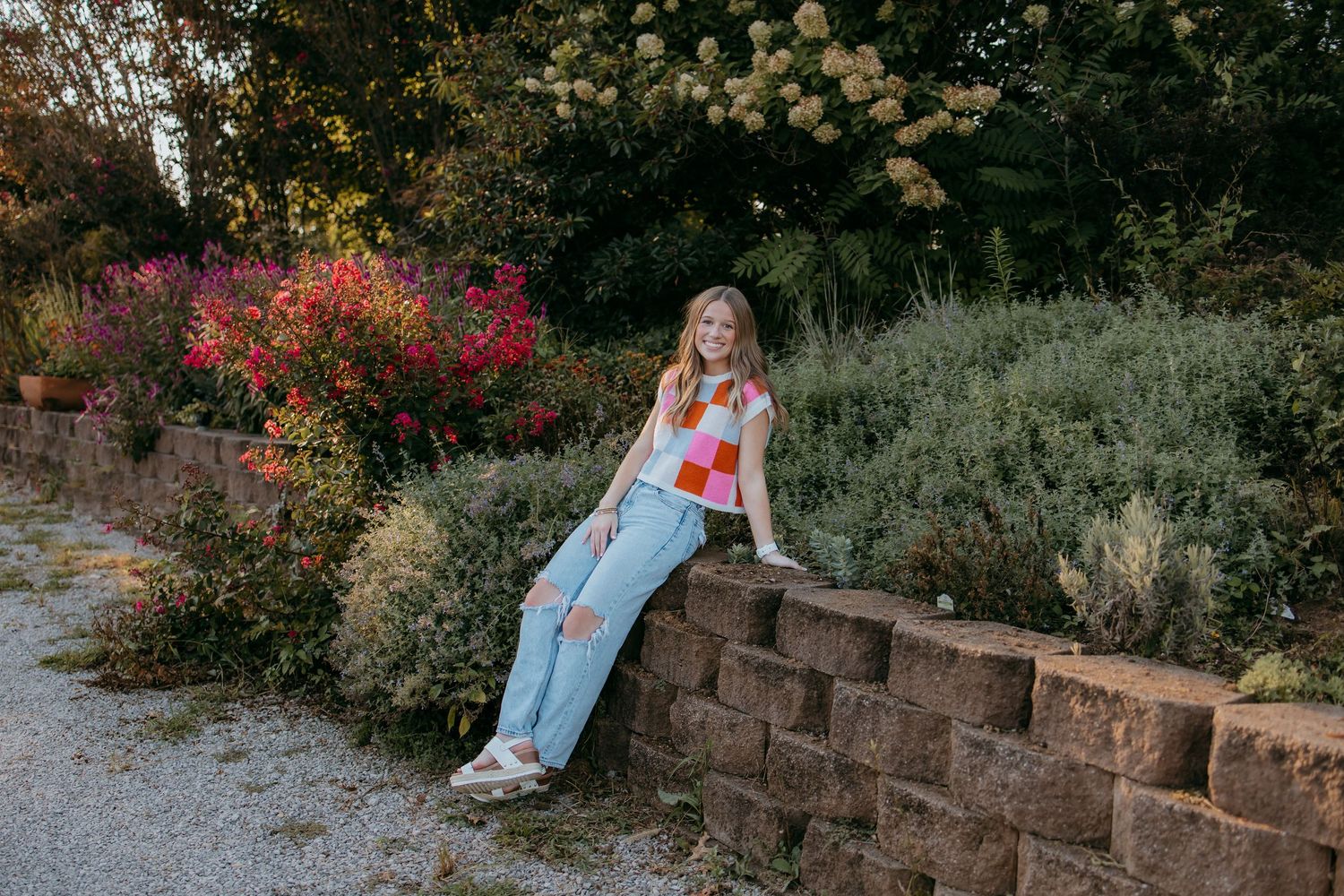 Senior in colorful checkered top and jeans sitting on a stone wall surrounded by vibrant garden flowers.