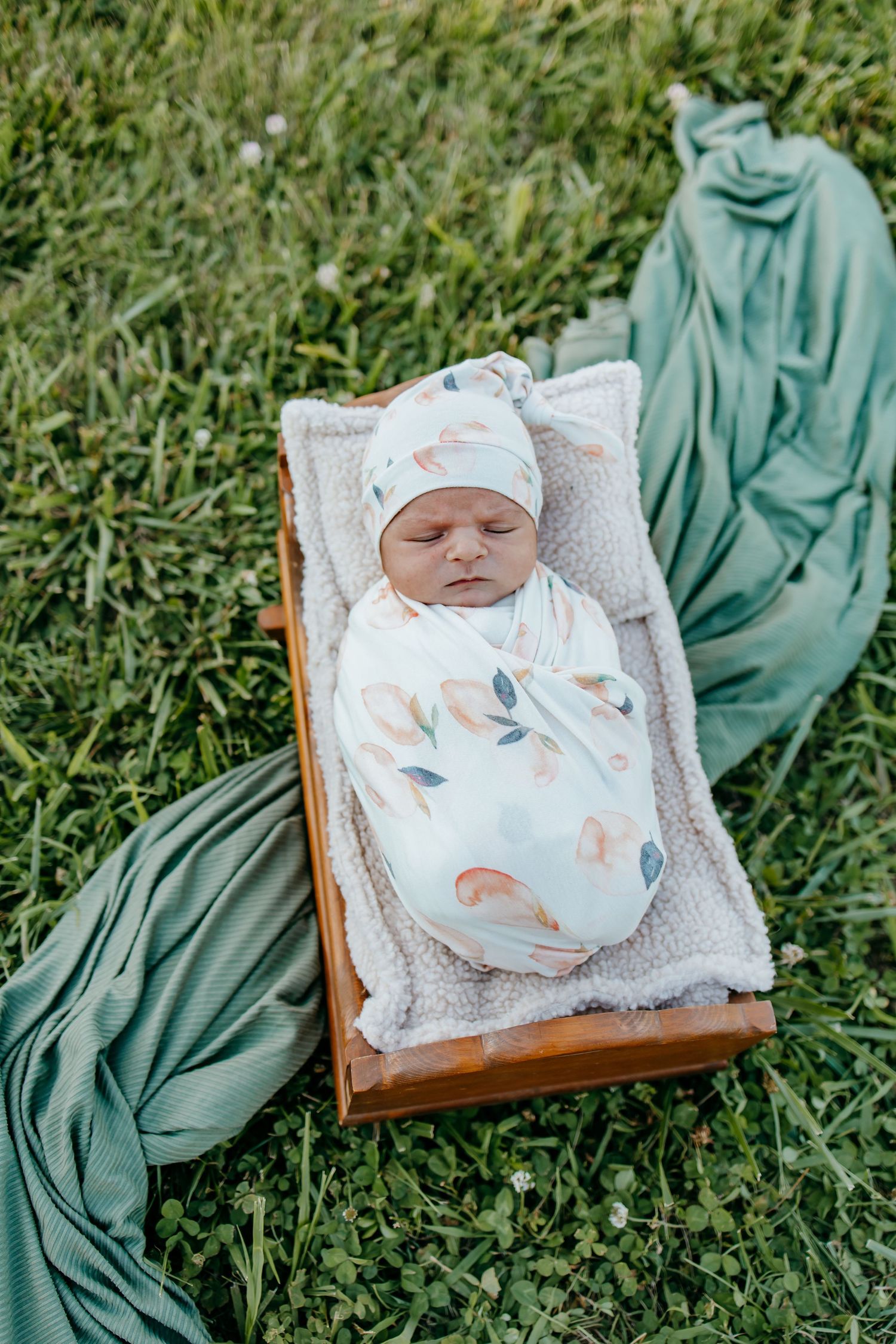 Newborn baby swaddled in floral wrap and knot hat, sleeping peacefully in a wooden crate on green grass.