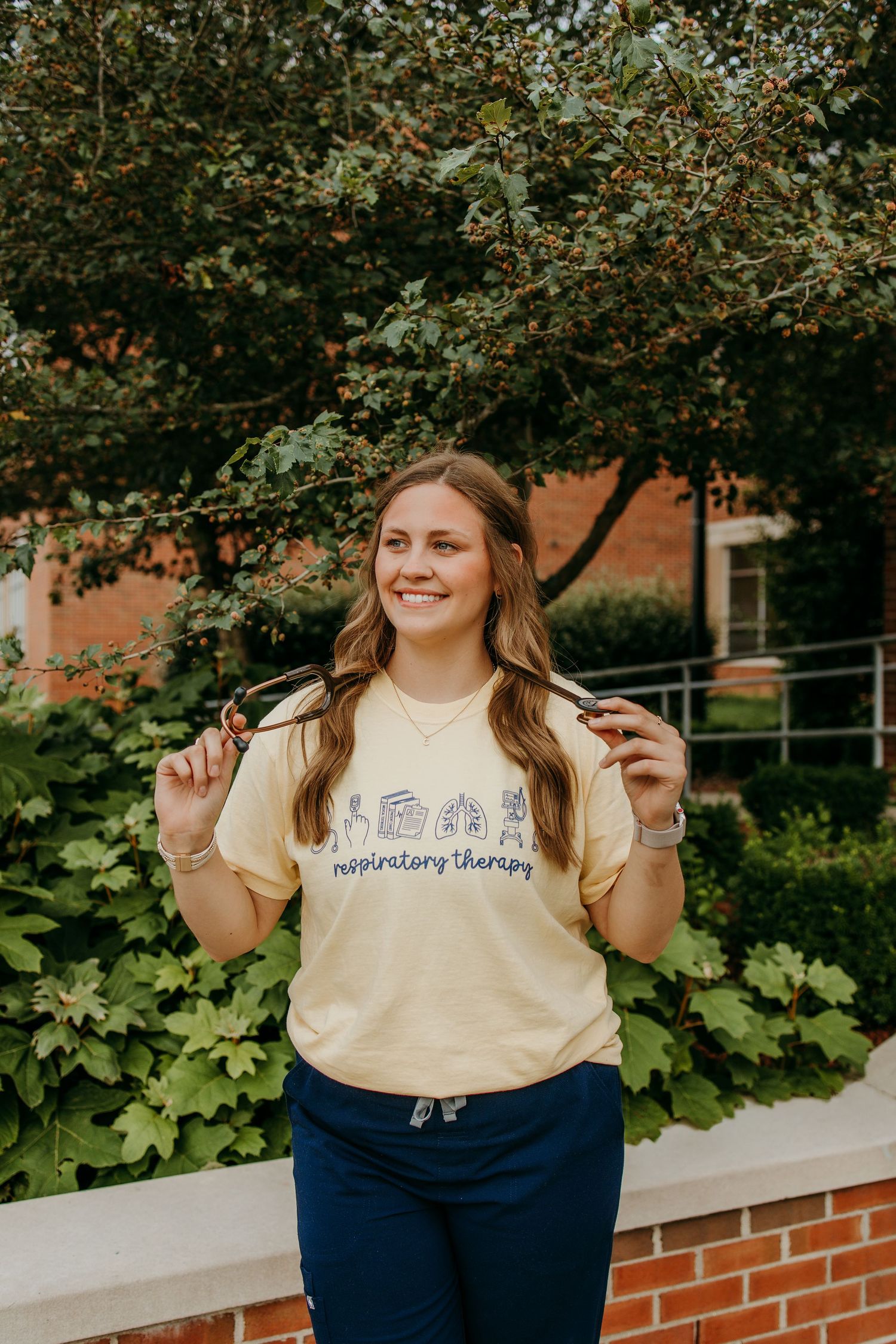 Smiling young woman in yellow respiratory therapy t-shirt and navy scrub pants posing outdoors near brick building.