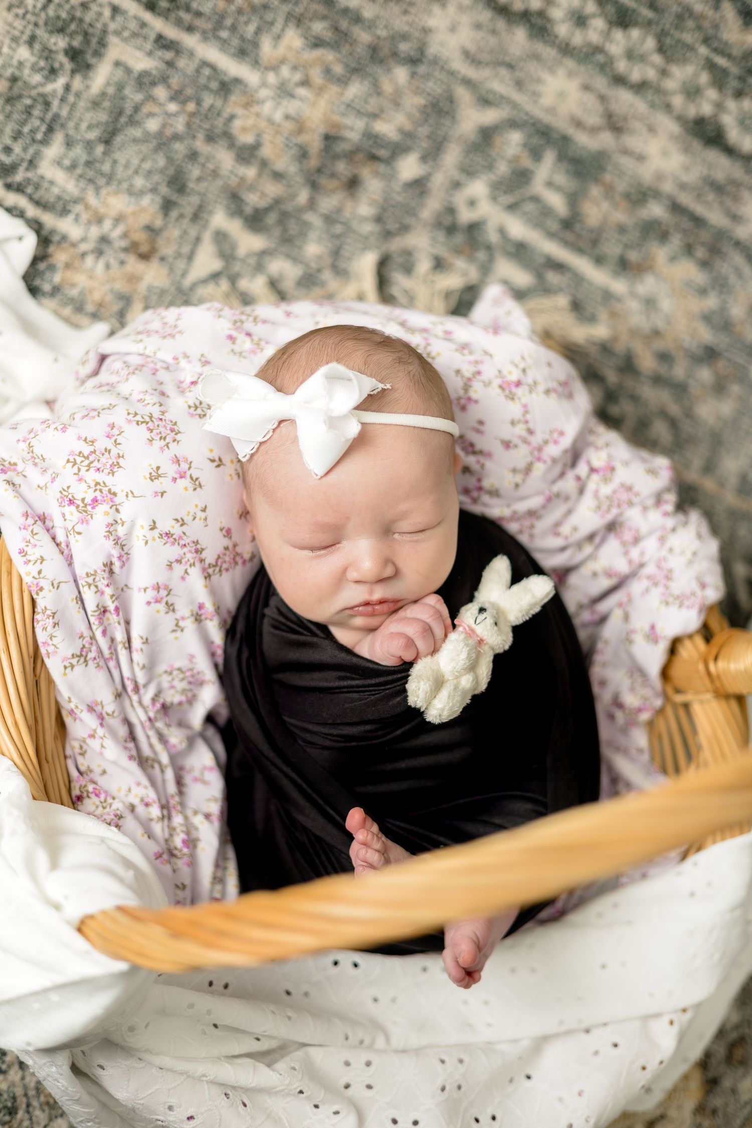 Sleeping newborn baby girl swaddled in black wrap with bunny toy, wearing white bow headband, resting in wicker basket.