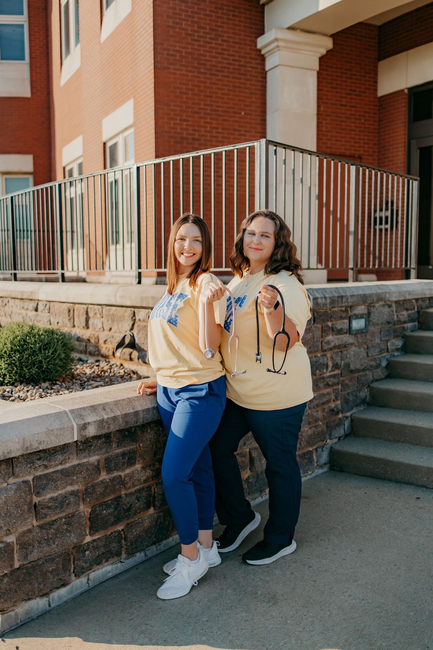 Two nursing grads in matching yellow medical-themed shirts pose smiling outside a brick building.