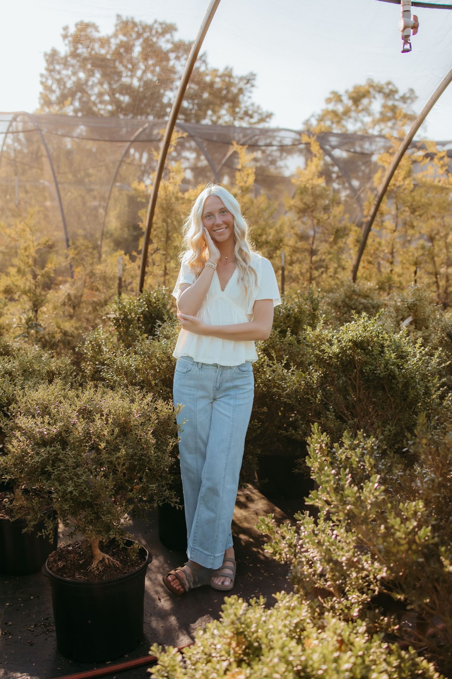 Smiling senior in white top and jeans standing among green shrubs in a sunlit greenhouse nursery.