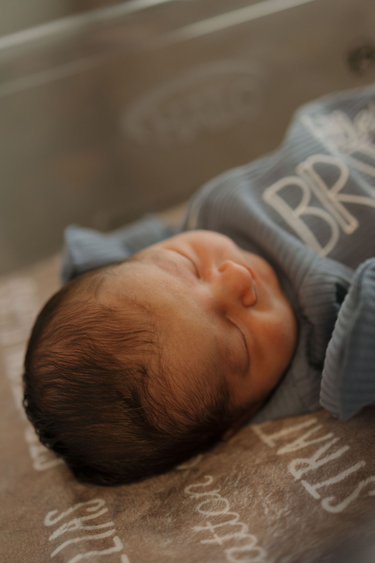 Sleeping newborn baby with dark hair in a blue sleeper in a hospital bassinet.