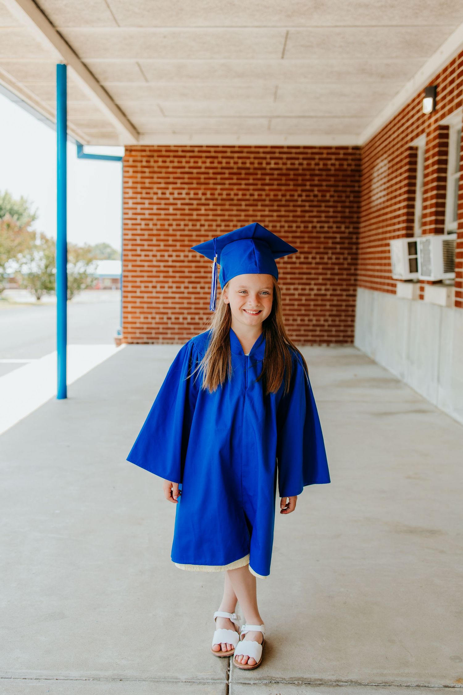 Young girl smiling in blue graduation cap and gown standing in school hallway corridor.