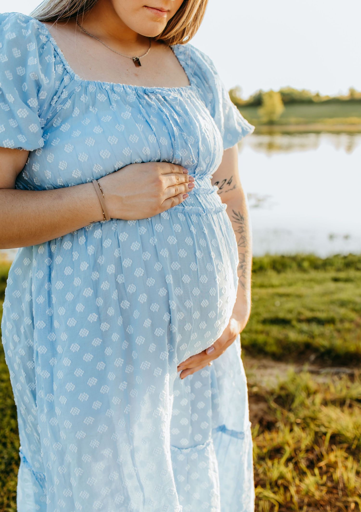 Pregnant woman in blue polka dot dress holding baby bump outdoors near a lake during golden hour.