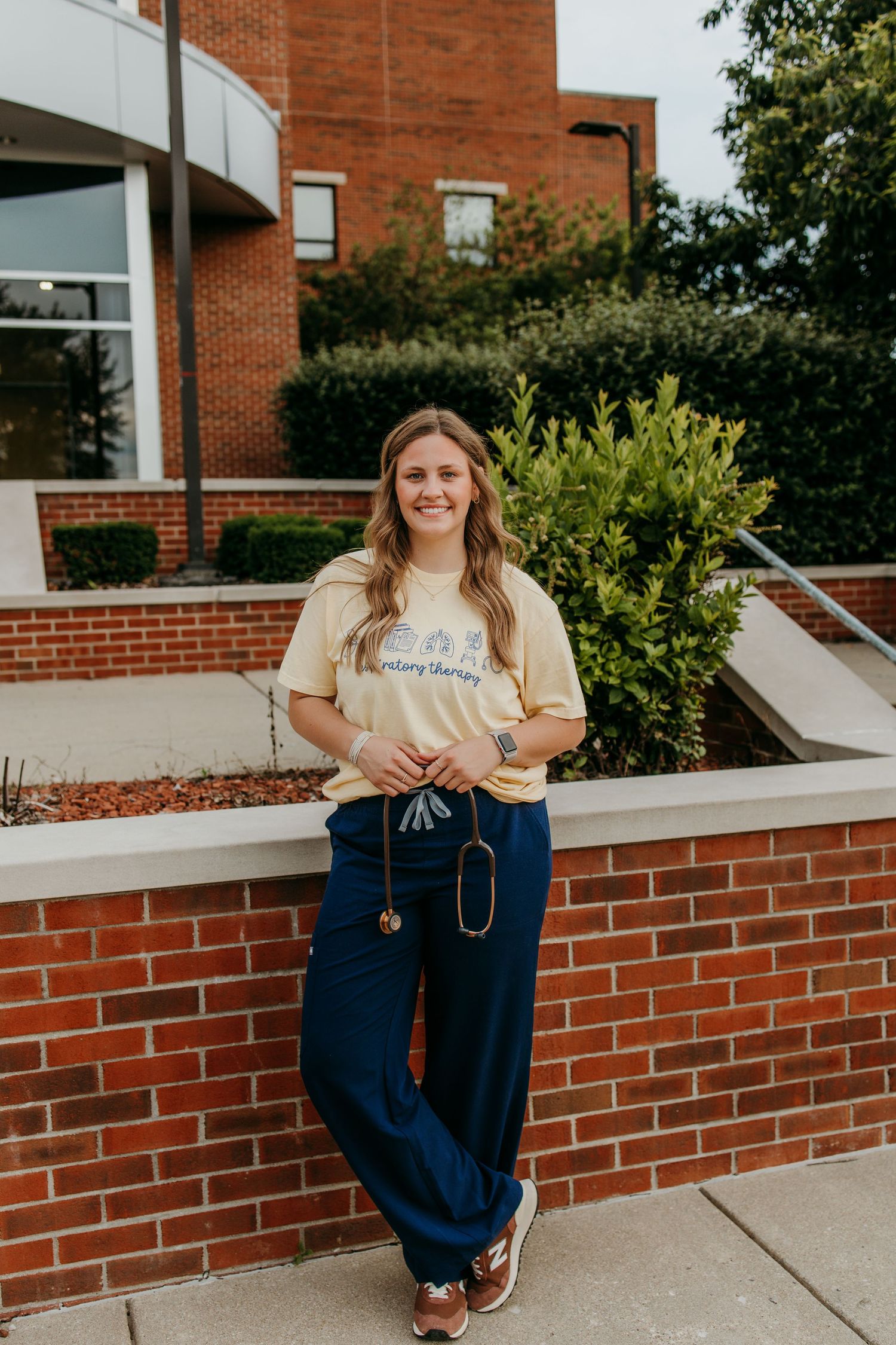 Smiling respiratory therapy student in yellow tee and navy scrubs holding stethoscope outside brick building.
