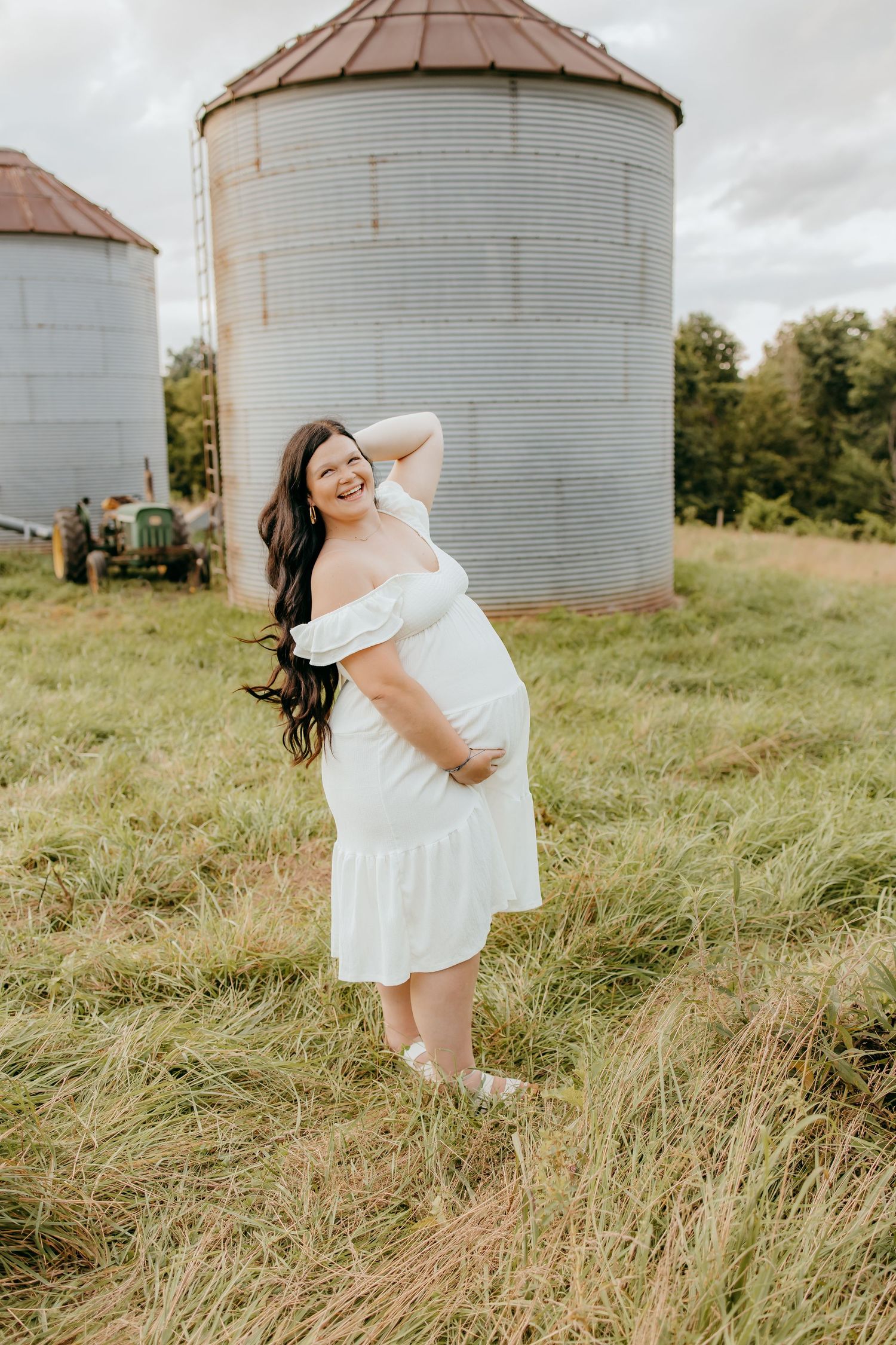 Pregnant woman in white off-shoulder dress posing in green field with grain silos in background.