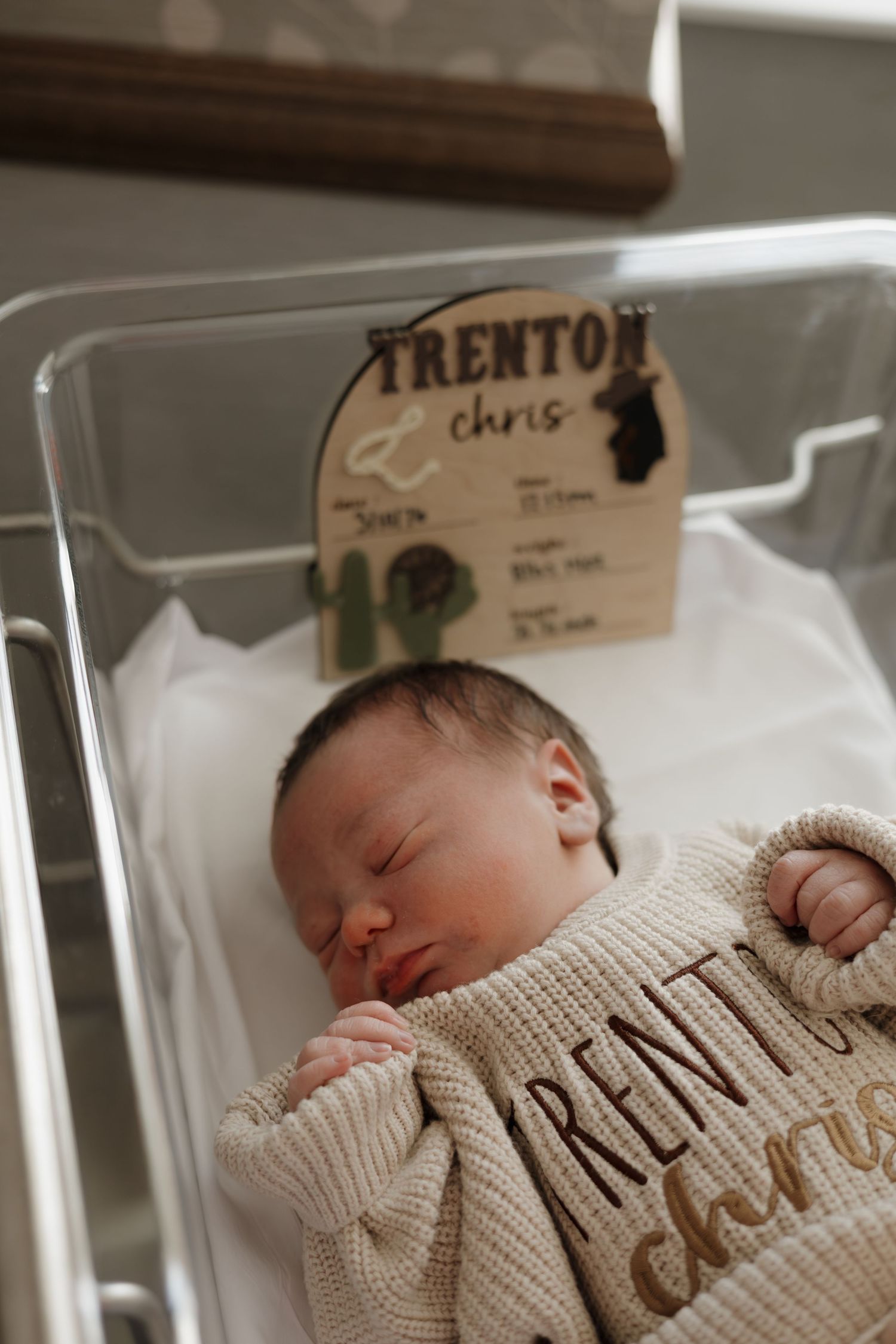 Newborn baby Trenton Chris sleeping peacefully in hospital bassinet, wrapped in personalized knit blanket with name card.