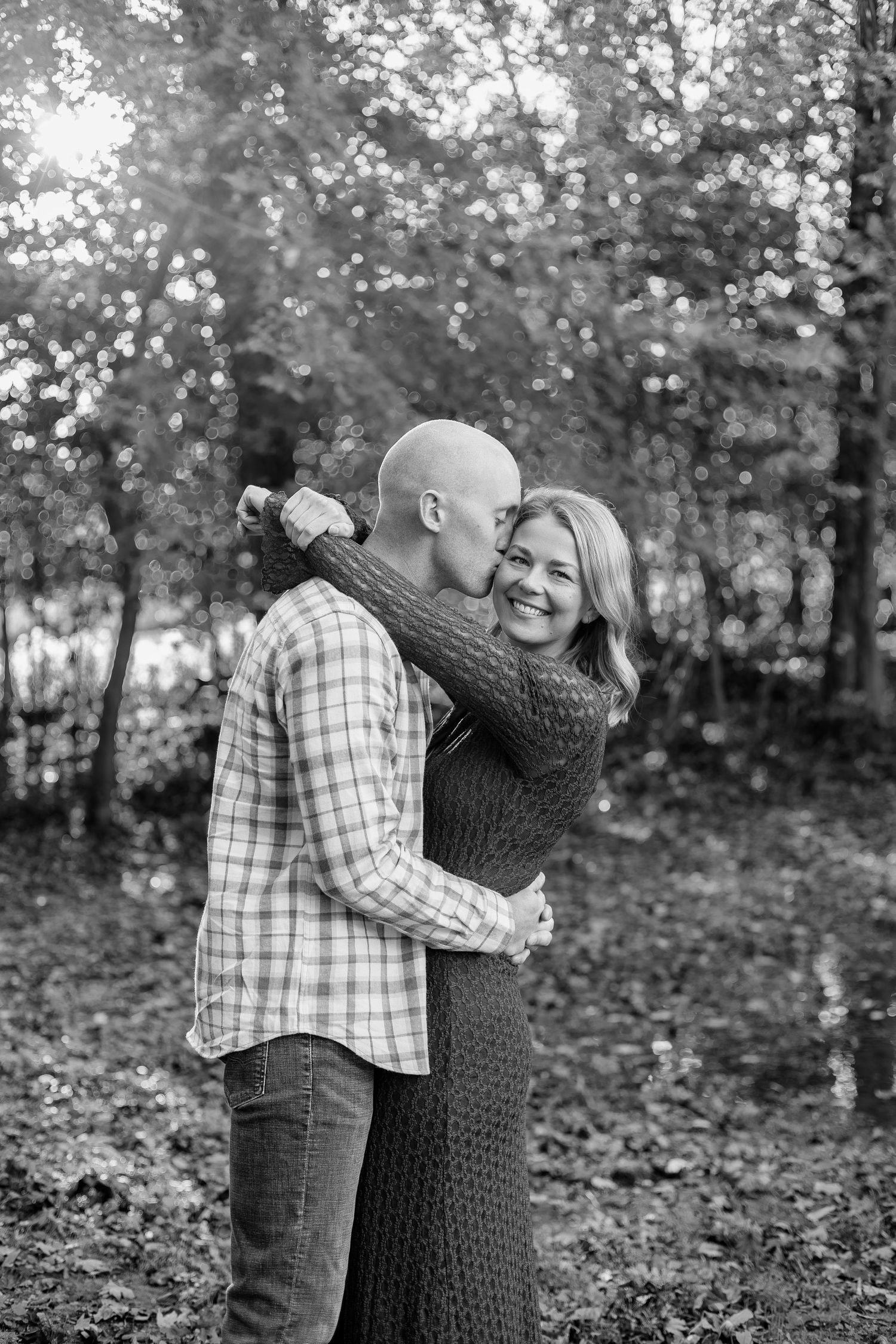 Black and white photo of a couple embracing in autumn woods, man kissing woman's cheek as she smiles warmly.