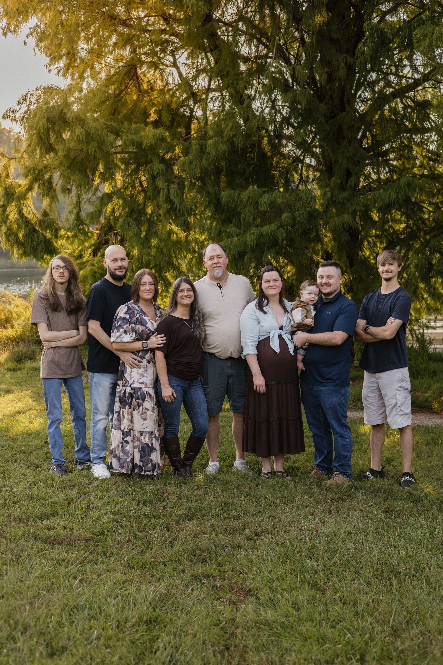 Family portrait of eight people standing outdoors on green grass beneath a large tree in golden hour light.