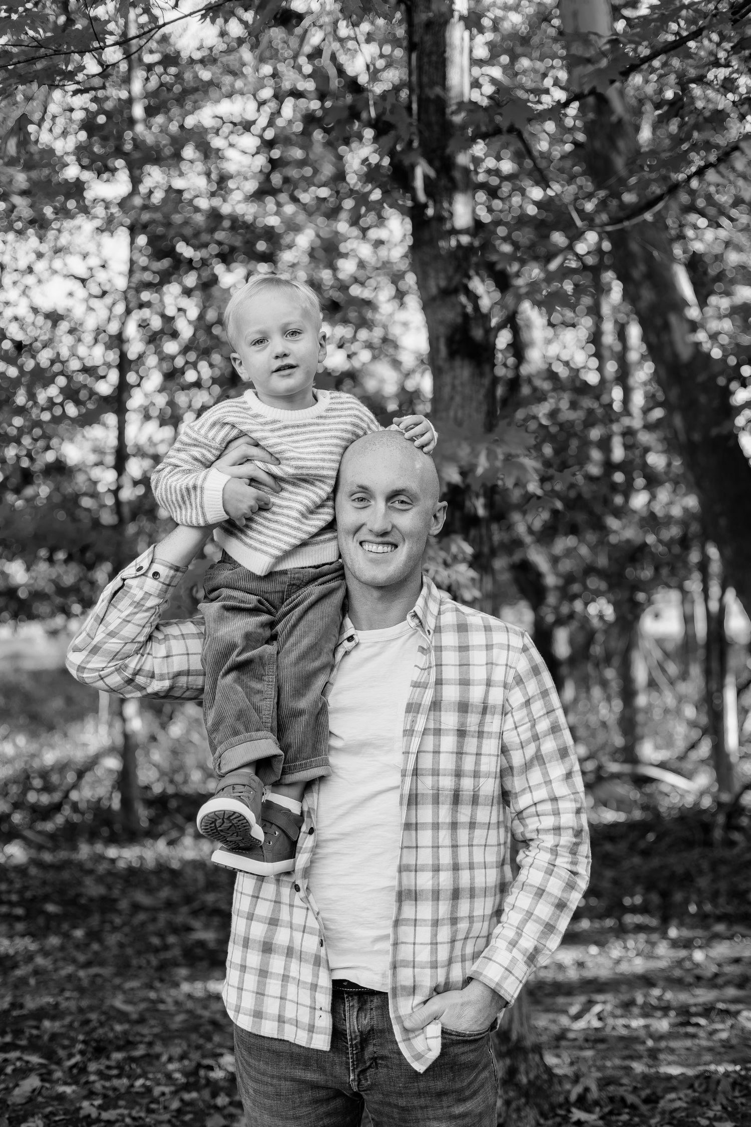 A smiling bald man carries a young toddler on his shoulders in a forest setting, captured in black and white.
