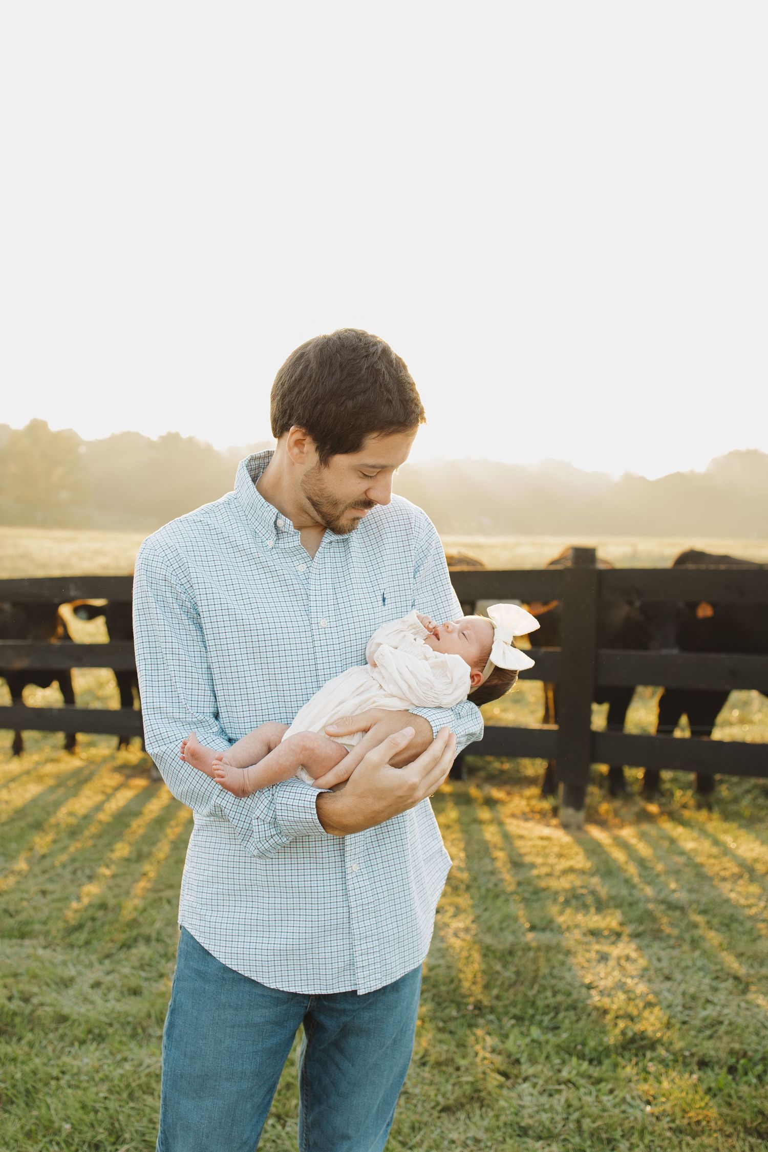 A man in a plaid shirt tenderly holds a newborn baby outdoors near a fence with cattle at golden hour.