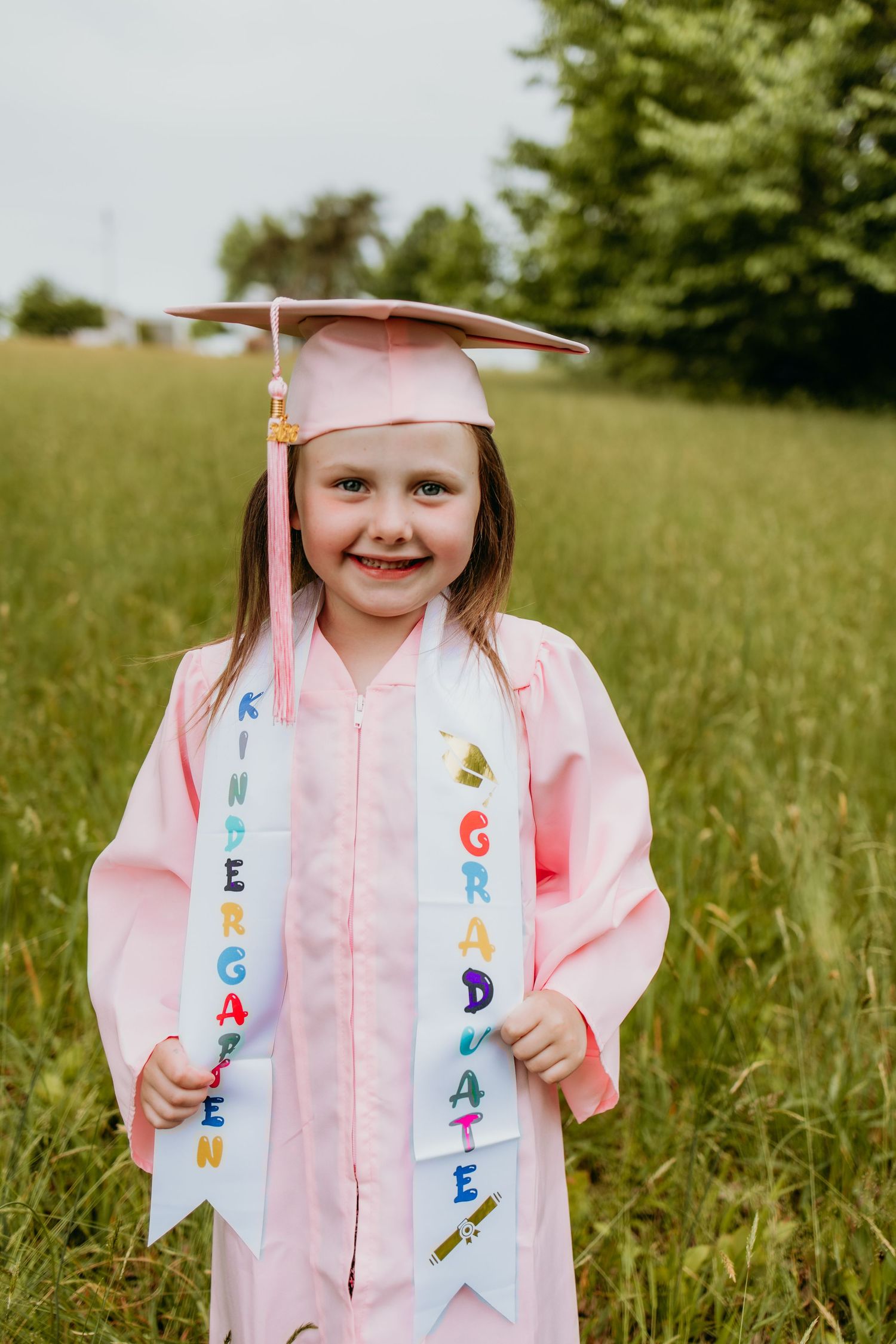 Young girl in pink kindergarten graduation cap and gown holding sashes outdoors in green field.