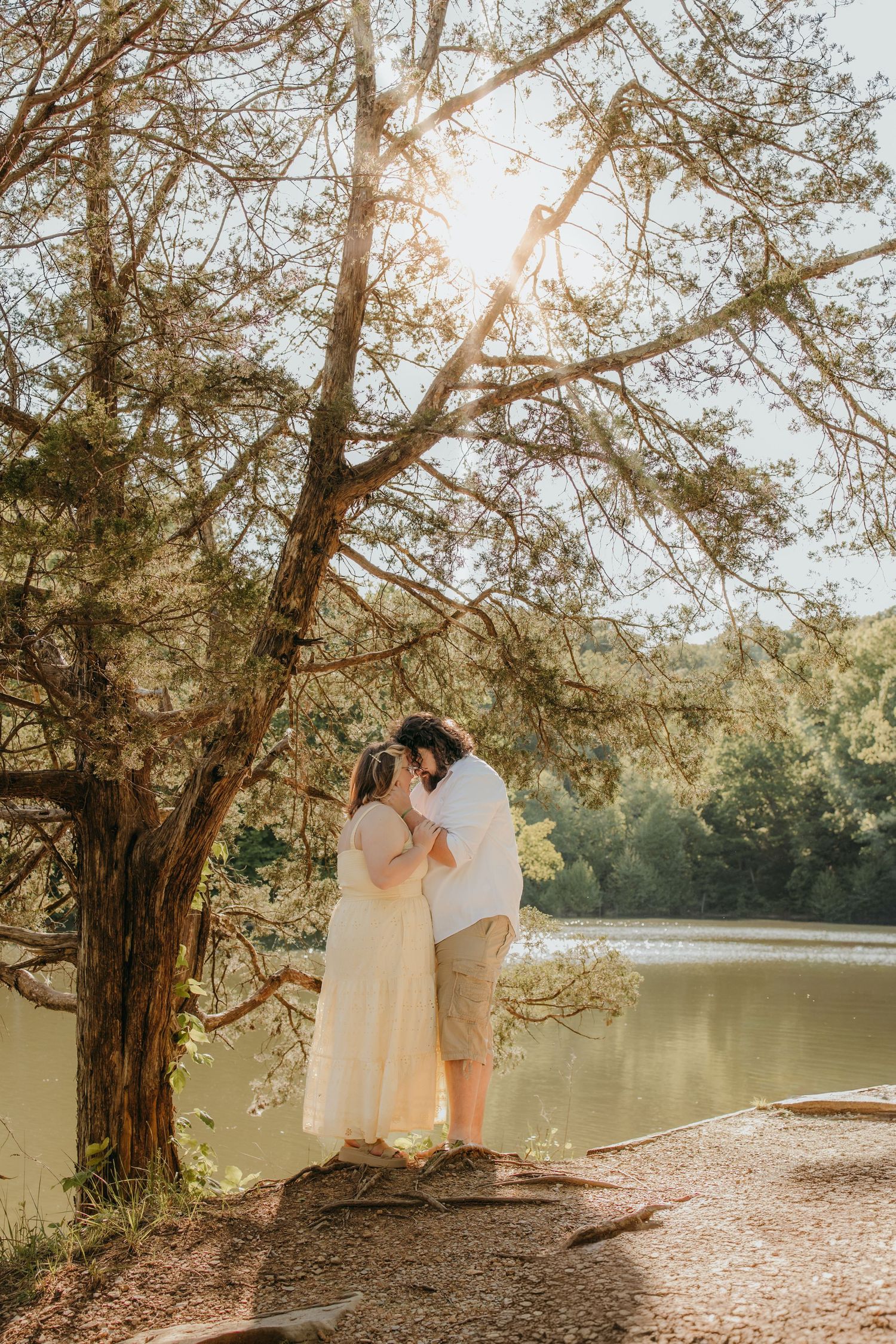 Romantic couple kissing under large tree by peaceful lake with golden sunlight streaming through branches.
