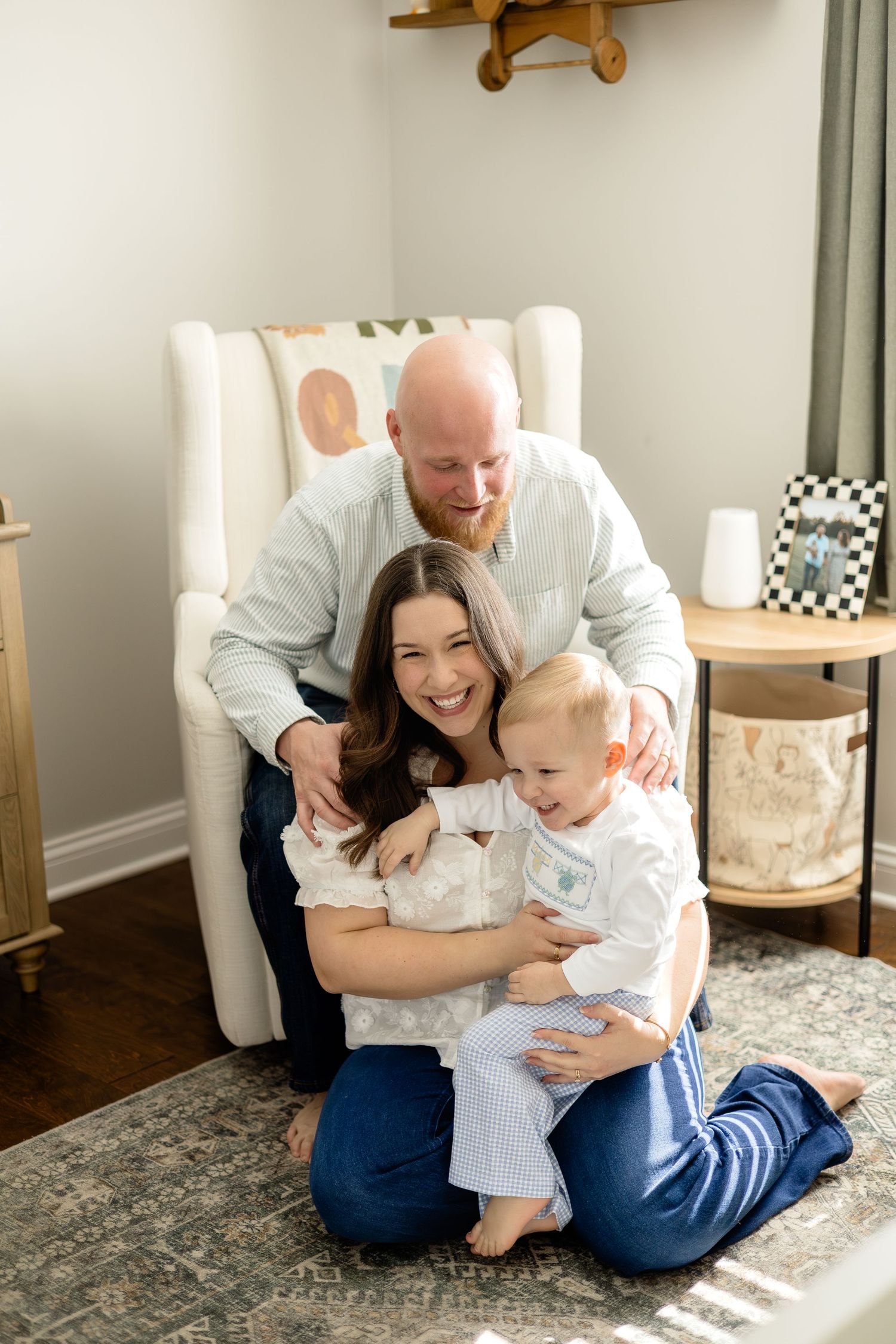 Happy family portrait with parents and toddler laughing together in a cozy nursery setting.