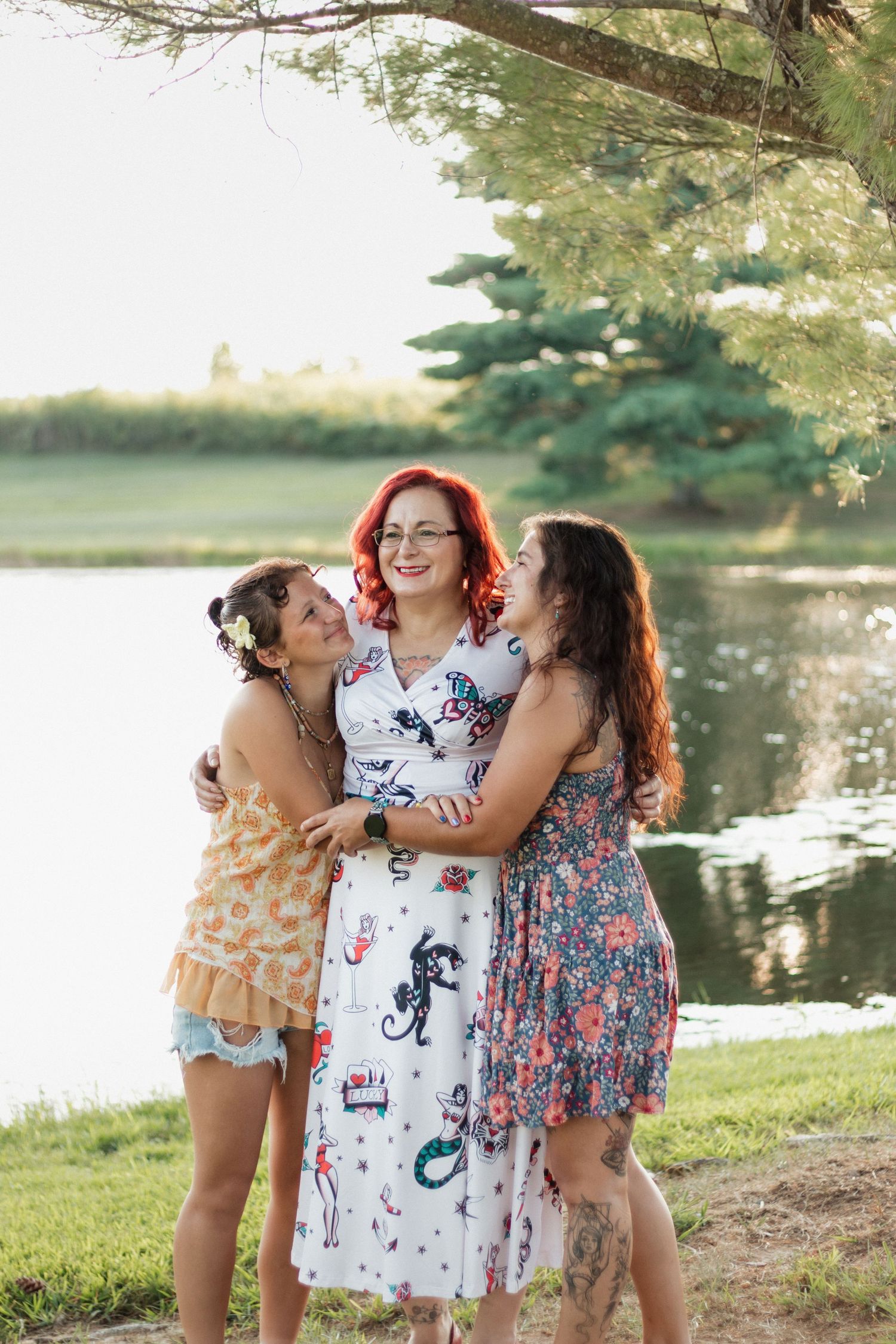 Three women in summer dresses embrace and smile near a serene lake surrounded by lush green trees.