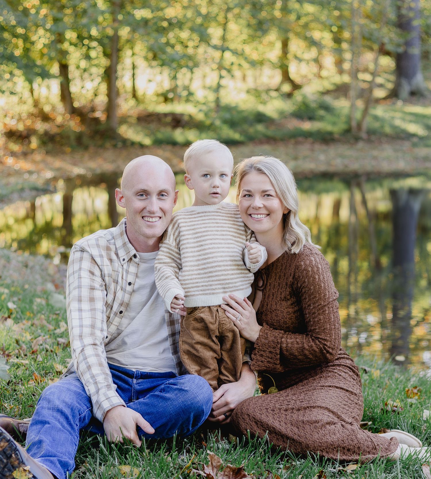 A smiling family of three poses outdoors by a pond, with parents sitting on grass holding their toddler son in autumn.