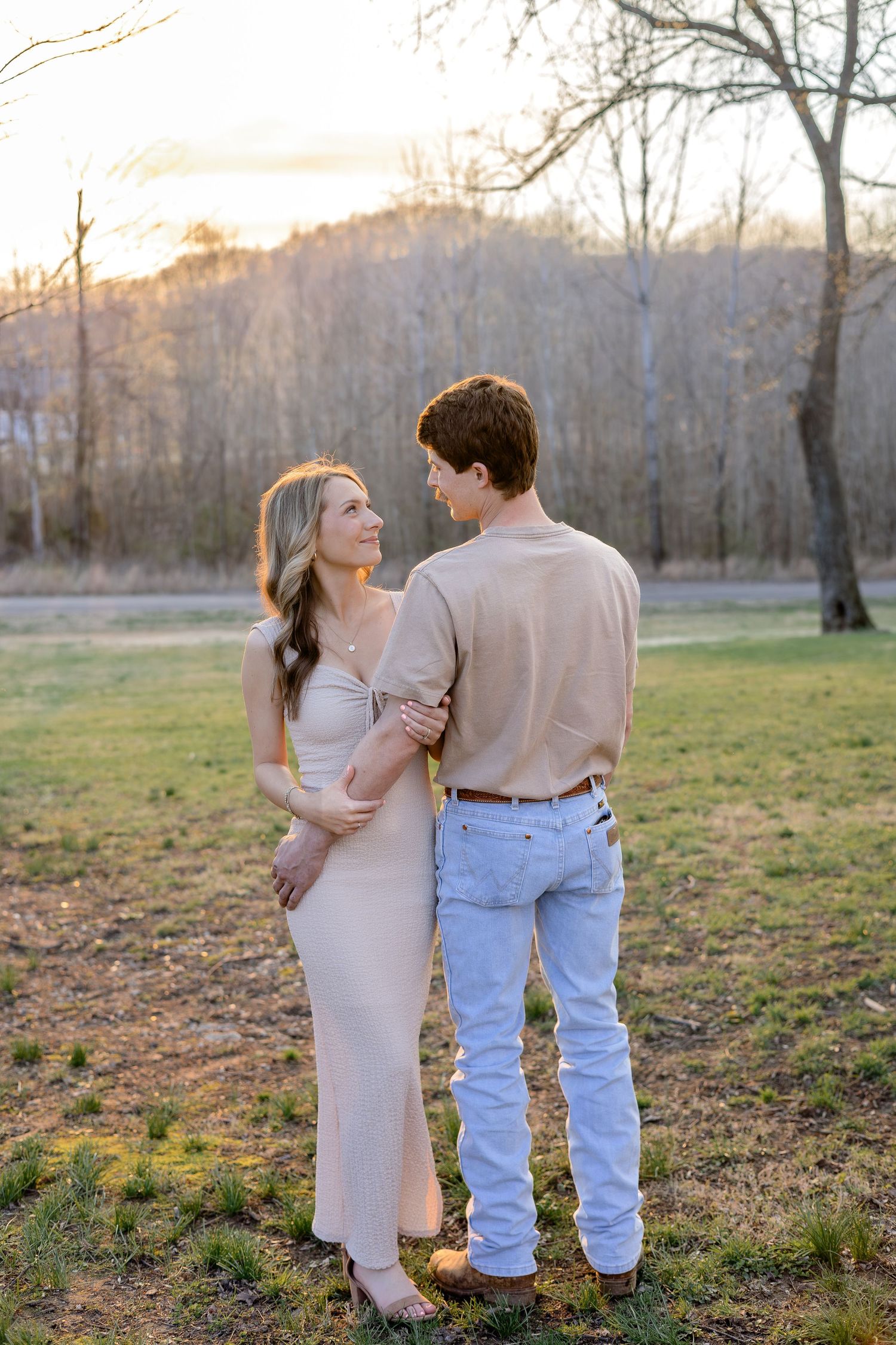 Couple in neutral tones sharing a tender moment in an open field during a warm golden sunset.