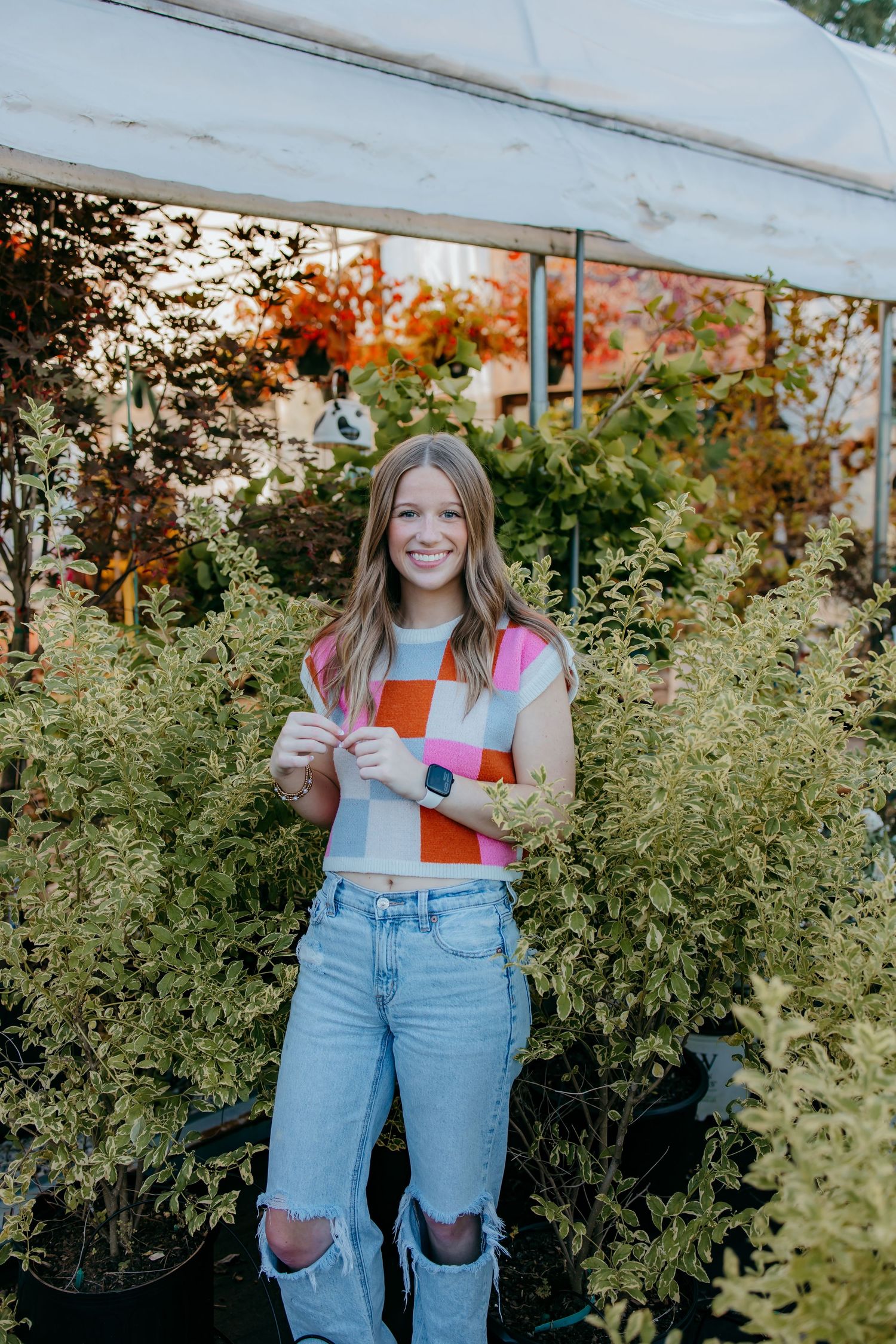 Senior in colorful checkered top and ripped jeans smiling among lush green plants at a garden nursery.