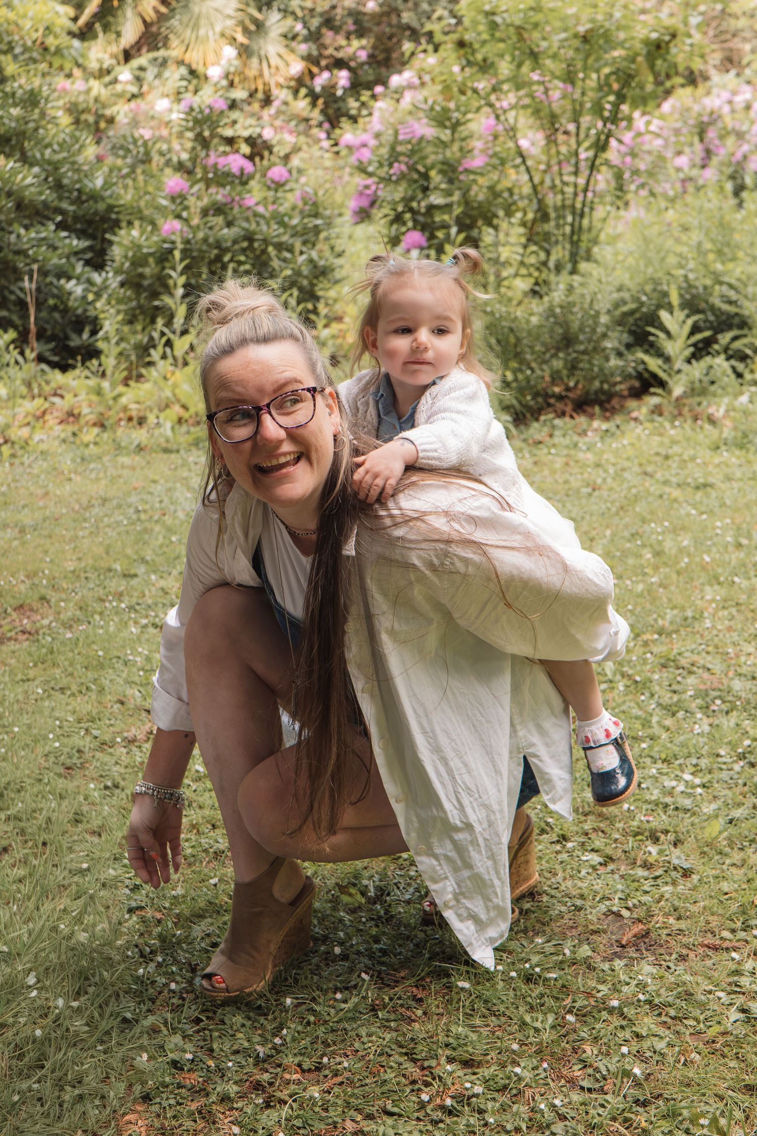 A laughing woman with glasses gives a toddler girl a piggyback ride in a lush green garden.