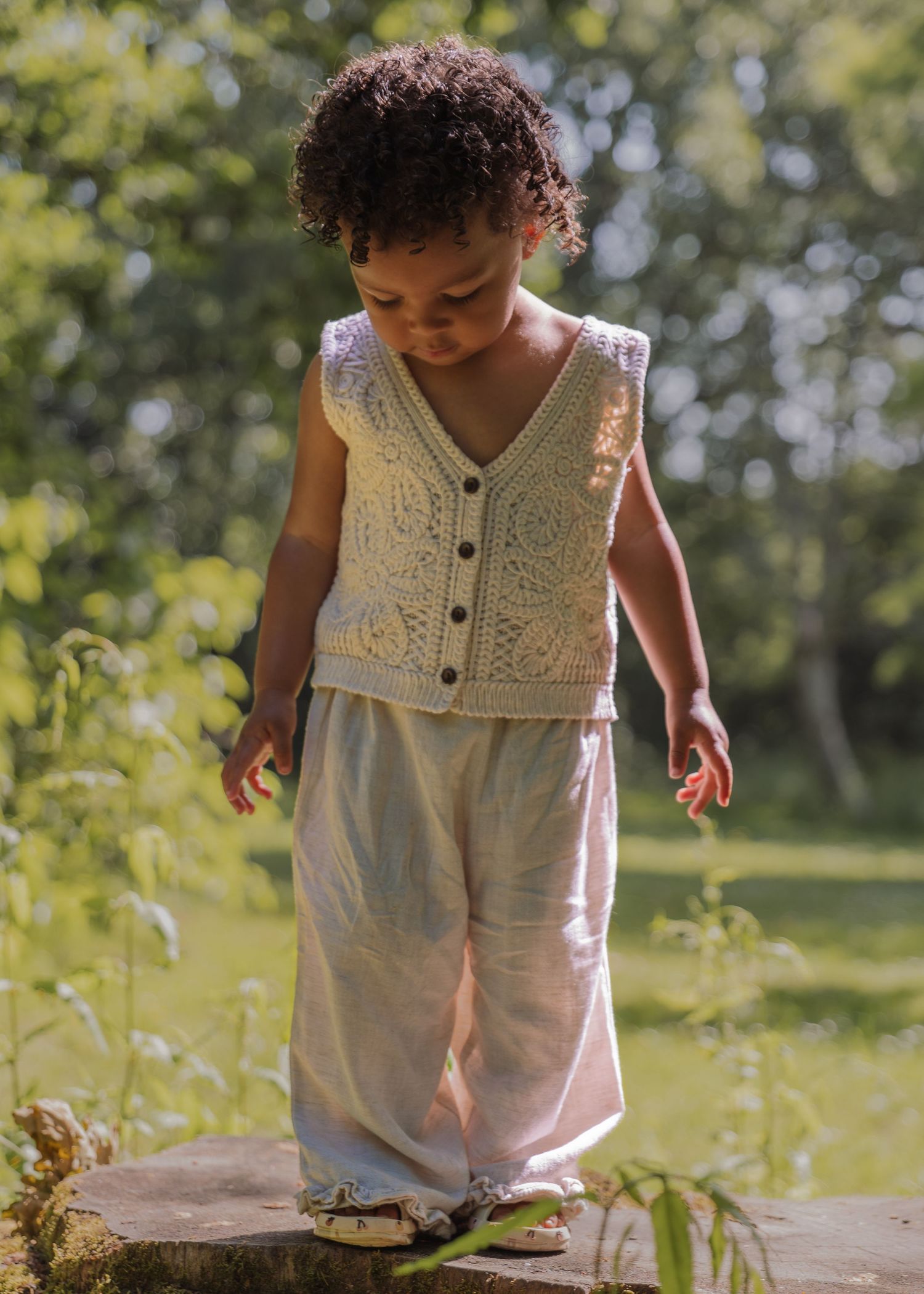 Toddler in cream lace vest and linen pants standing outdoors in dappled sunlight.