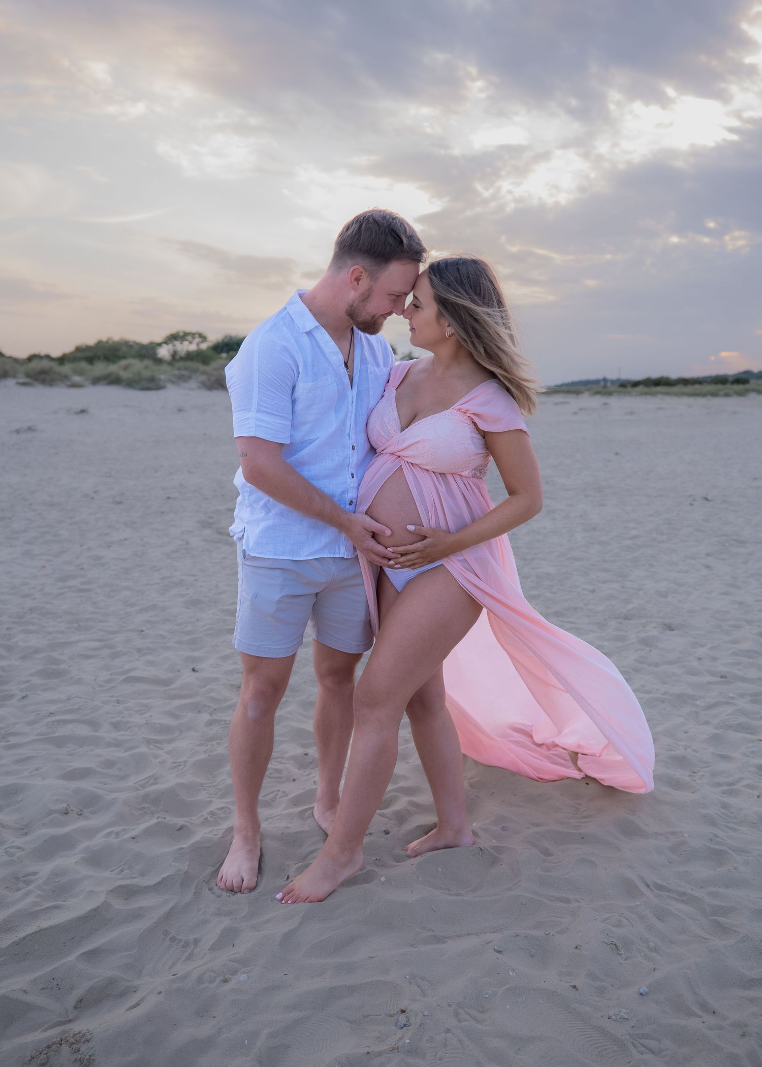 Couple sharing a tender moment on a sandy beach at sunset, woman in flowing pink maternity dress cradling her baby bump.