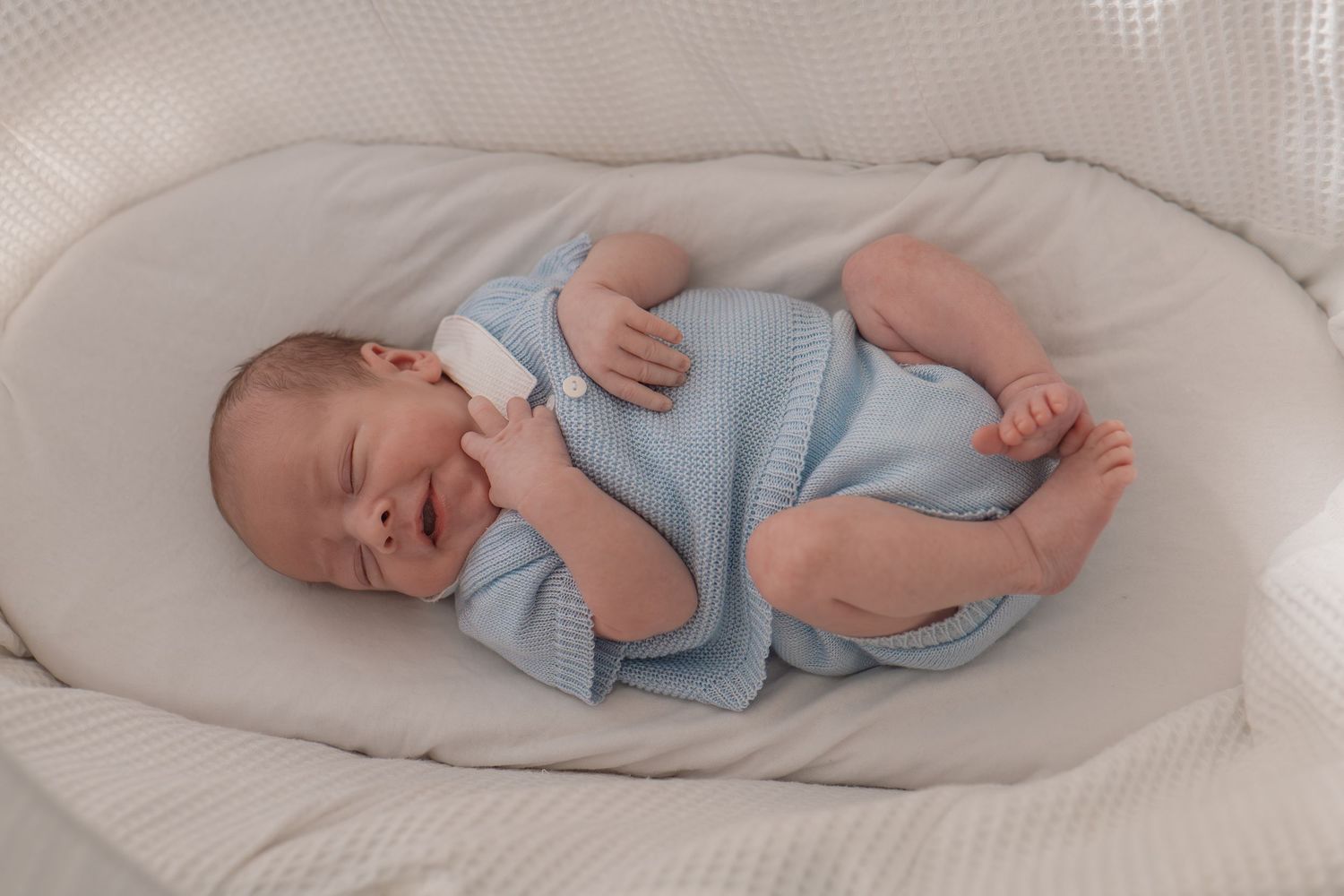 Newborn baby in blue knit outfit sleeping peacefully in a white cushioned bassinet.