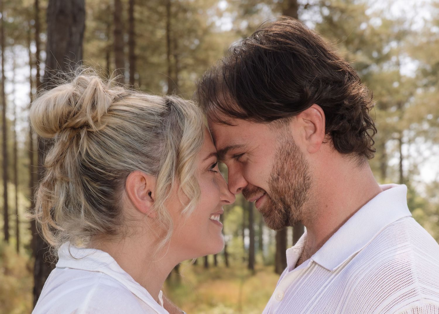 A couple in white shirts share a romantic moment, nearly kissing, surrounded by tall trees in a forest setting.