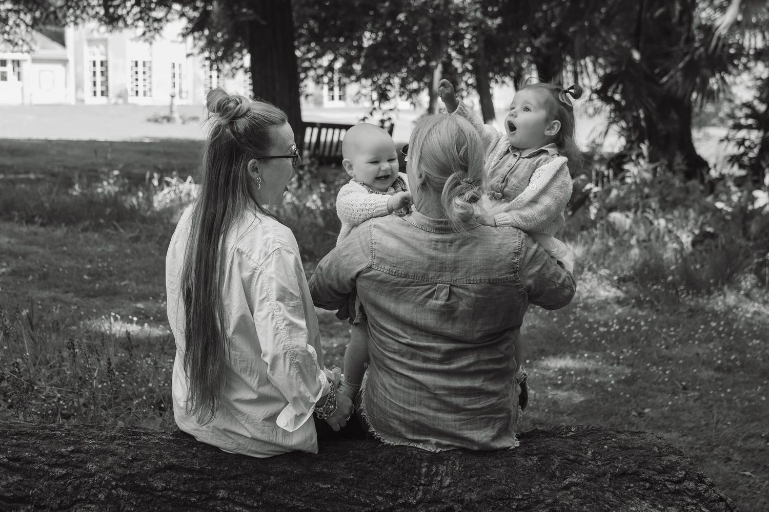 Black and white photo of two women sitting outdoors holding two happy toddlers in a lush garden setting.