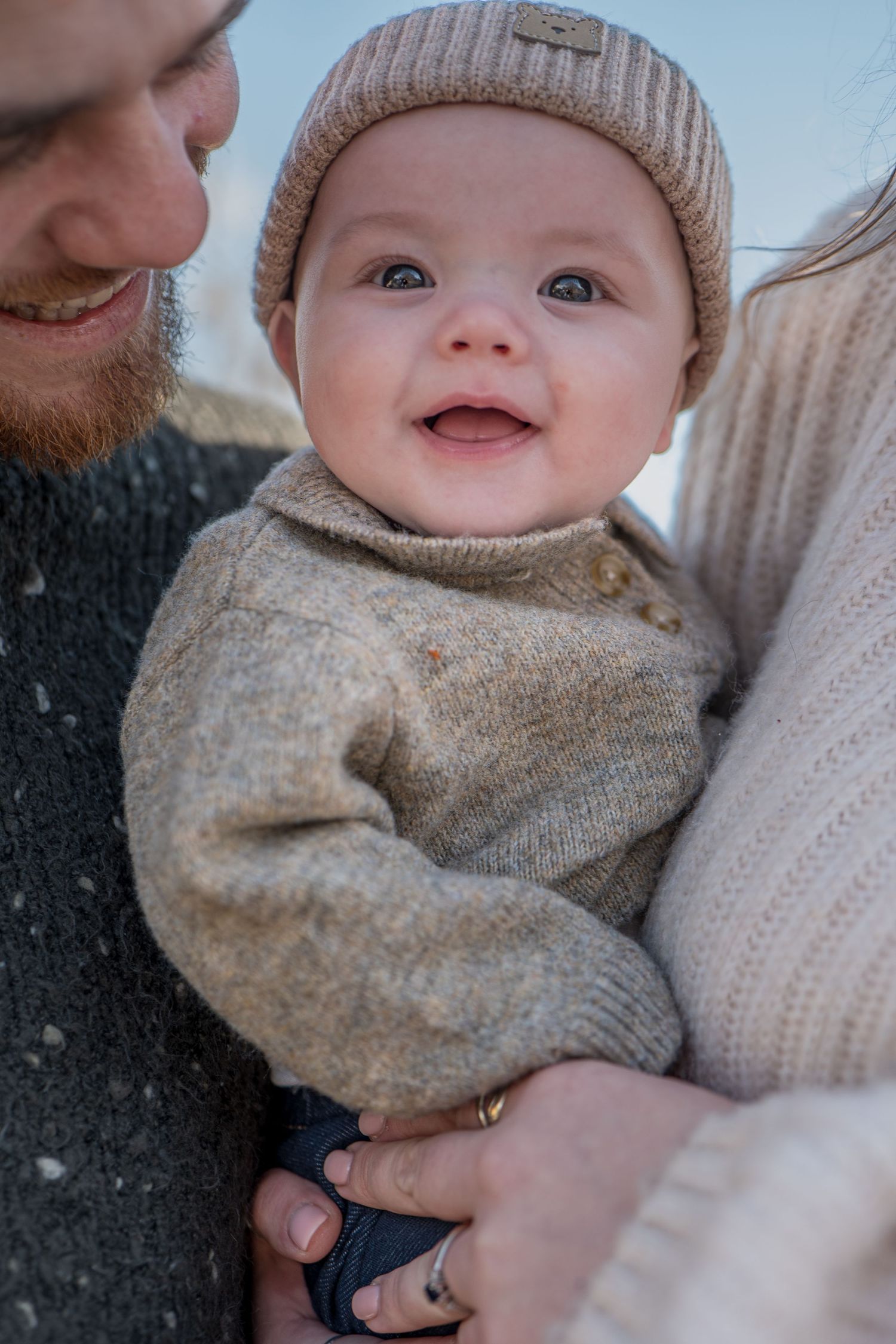 Happy baby in beanie hat smiling while being held by parents outdoors.