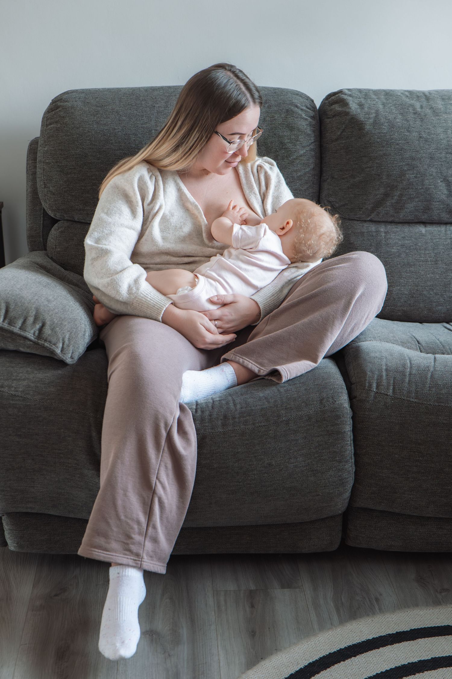 Young mother with glasses breastfeeding newborn baby while sitting comfortably on gray sofa at home.