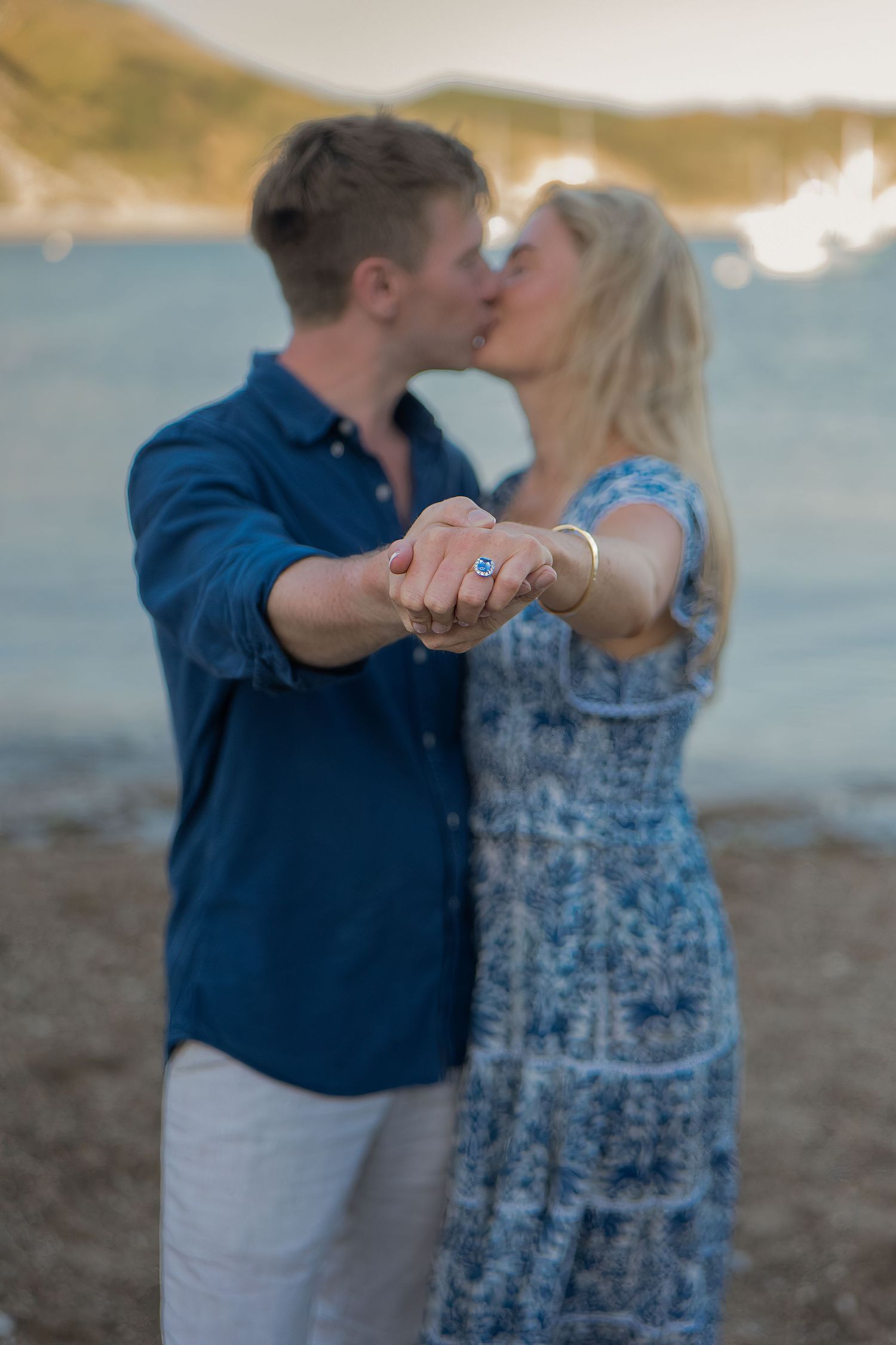 Engaged couple kissing on a beach while showing off their engagement ring to the camera.