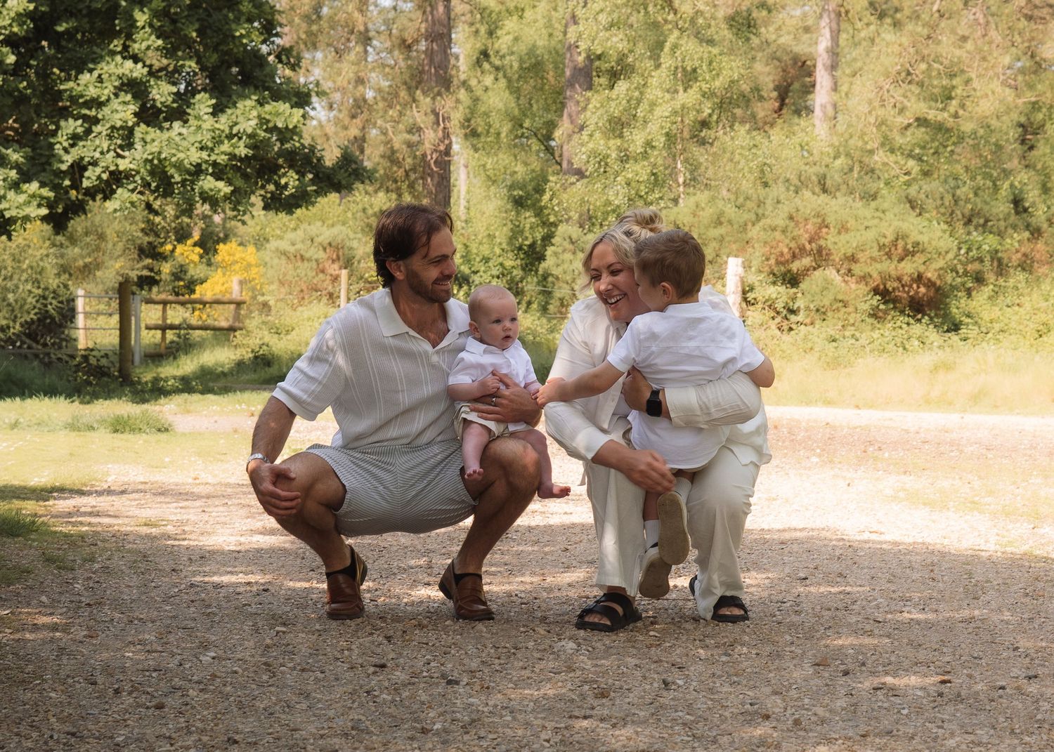 Family of four in white outfits enjoying a sunny outdoor moment on a dirt path surrounded by lush green trees.