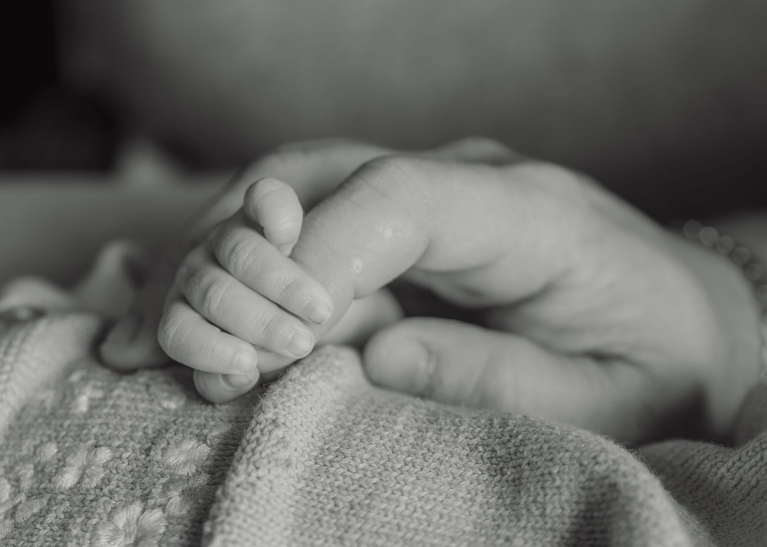 Black and white photo of a newborn baby's tiny hand gently gripping an adult's finger on a soft blanket.