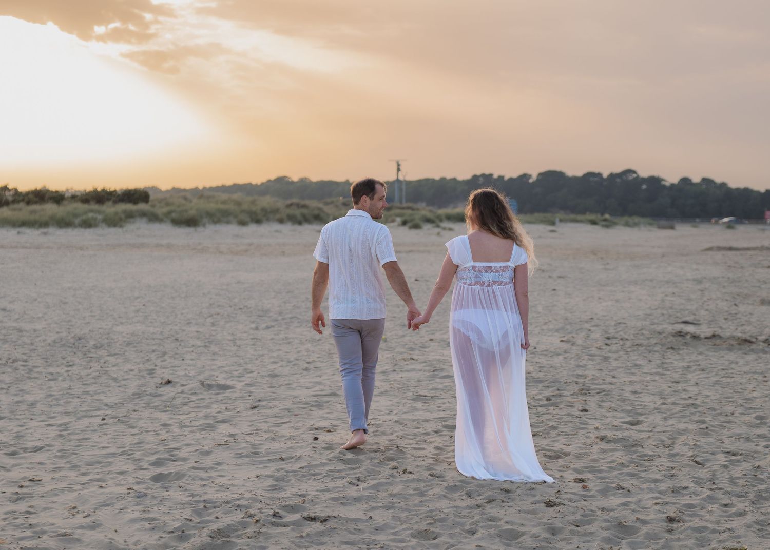 Couple holding hands walking on sandy beach at sunset, woman in white dress, man in white shirt and gray pants.