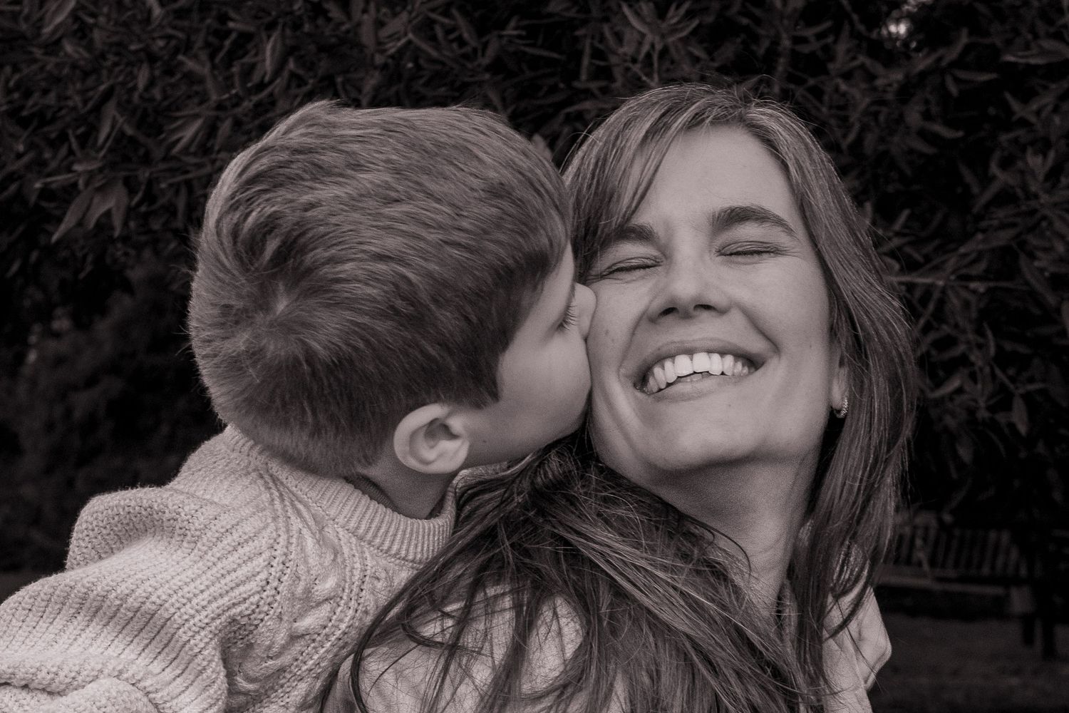 Black and white photo of a young boy kissing a smiling woman on the cheek outdoors.
