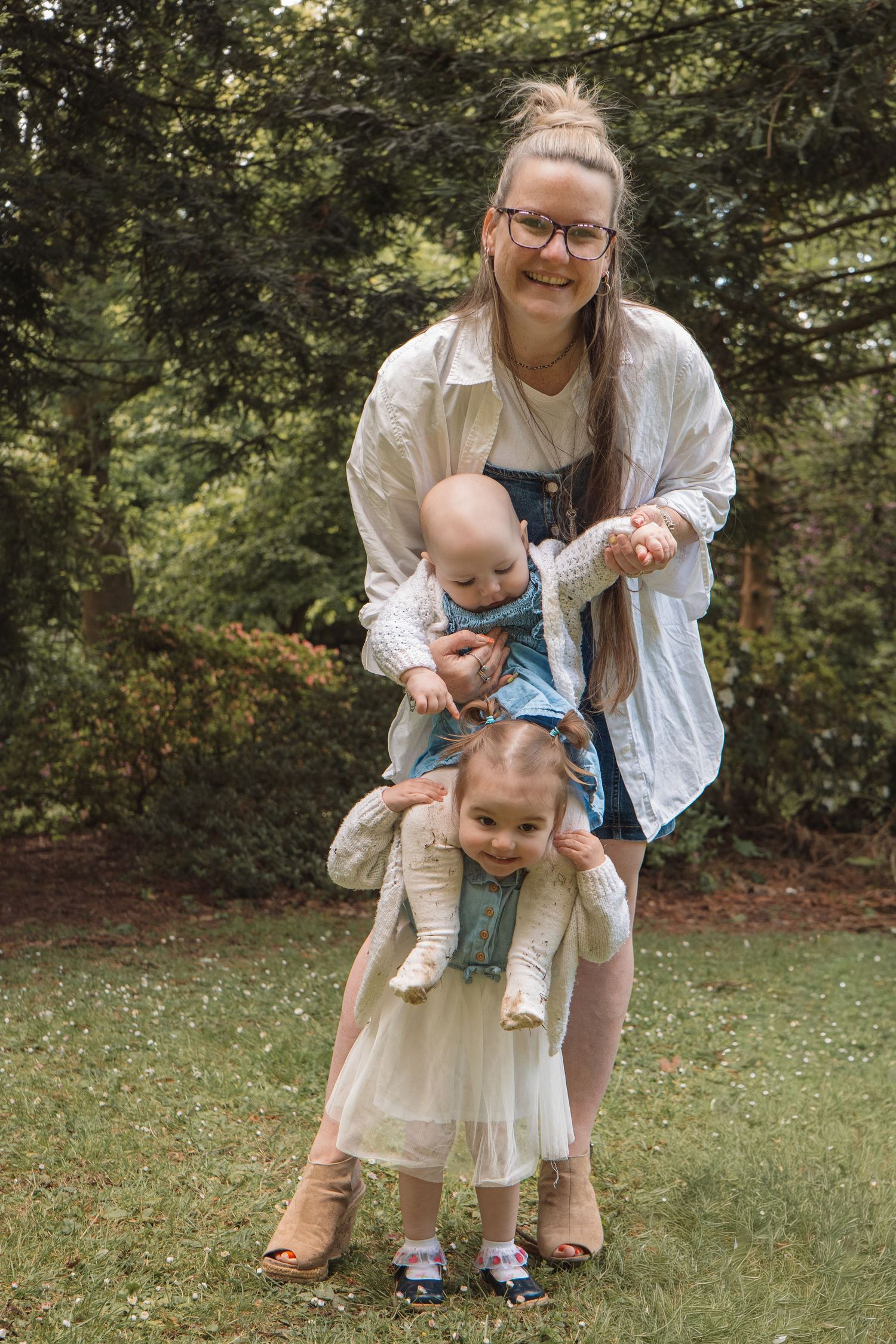 A smiling woman with glasses holds a baby and a toddler girl outdoors in a green park setting.