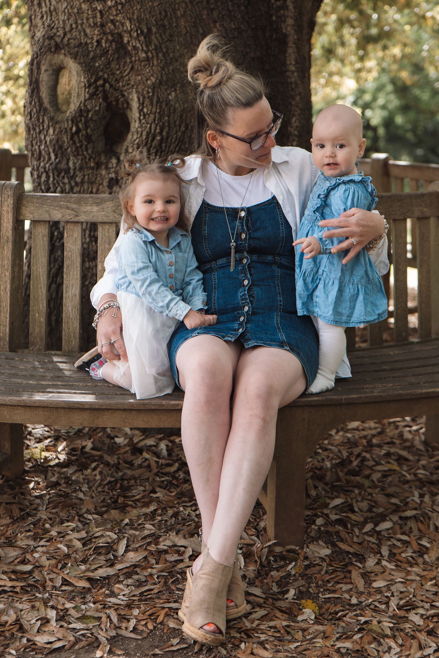 A mother in a denim dress sits on a wooden bench holding two young daughters in matching blue outfits outdoors.