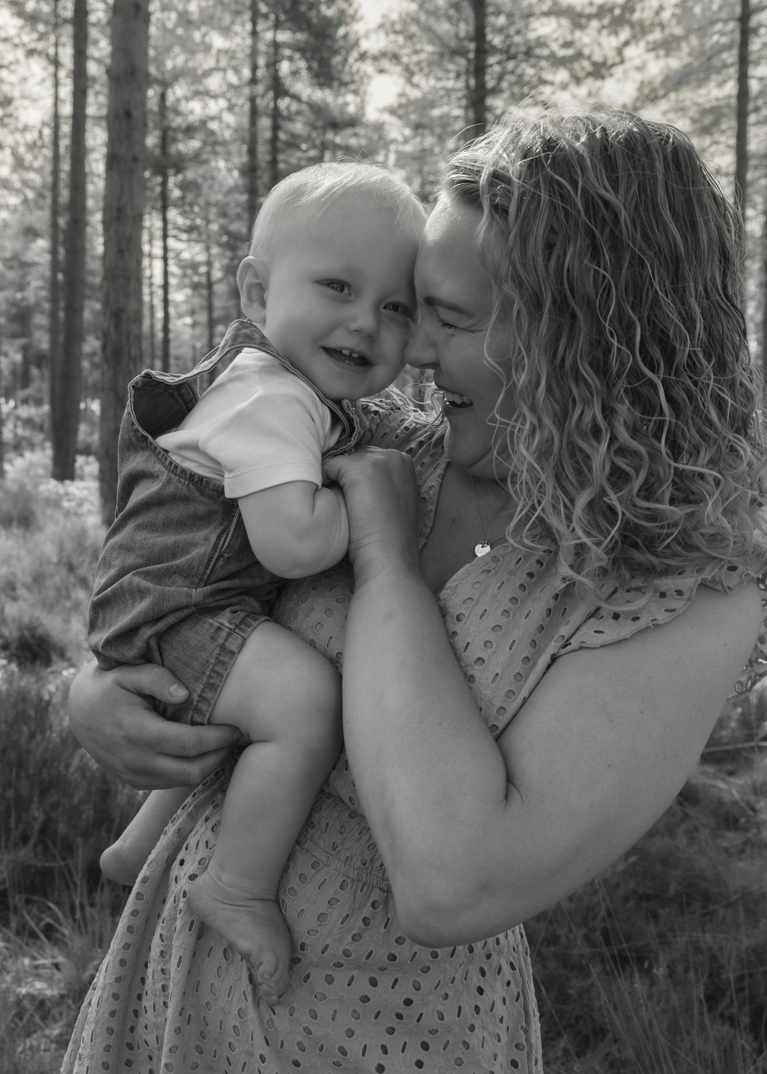 Black and white photo of a curly-haired woman holding and nuzzling a smiling baby outdoors among tall trees.