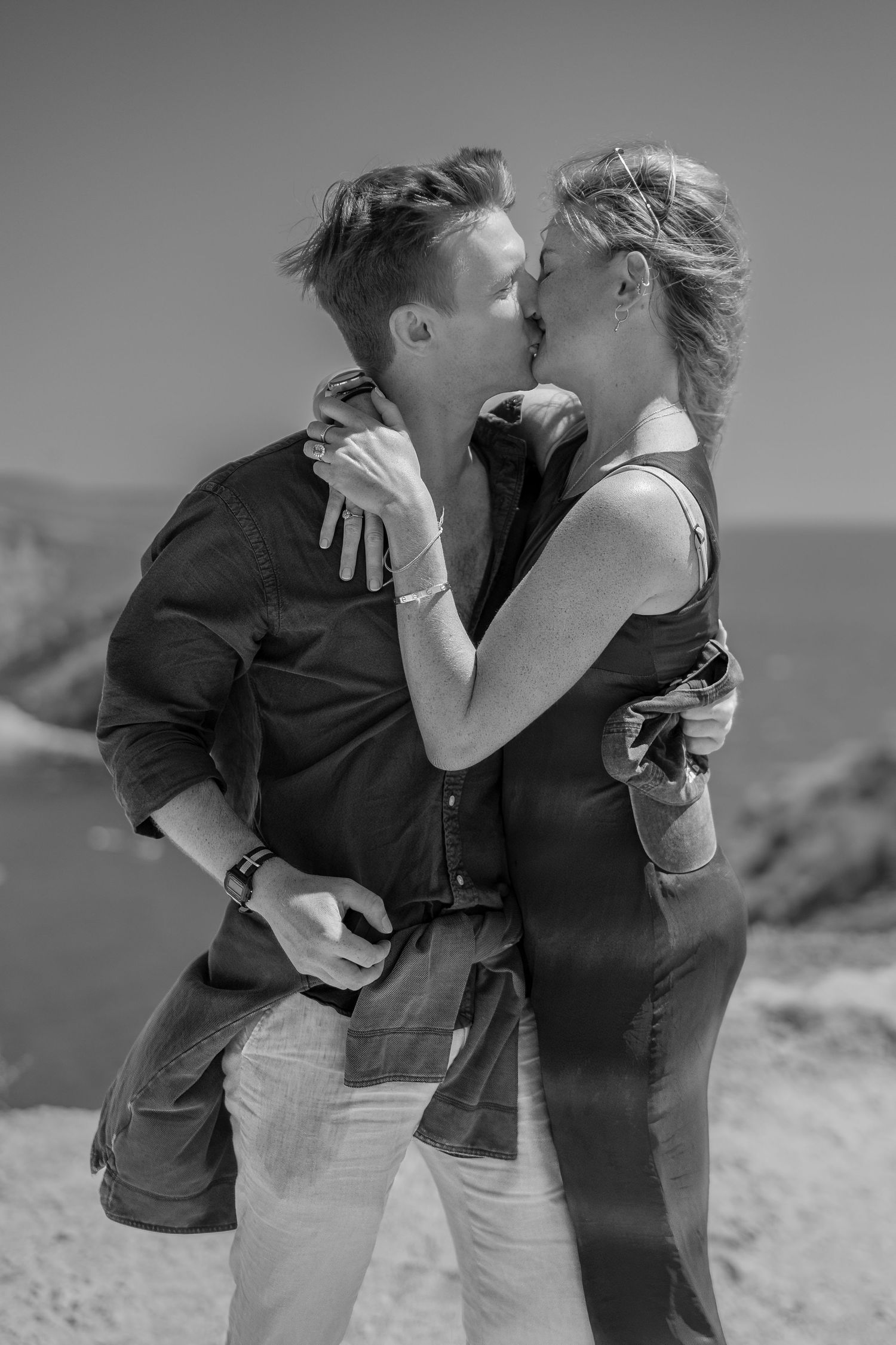 Black and white photo of a romantic couple kissing outdoors near a coastal cliff on a sunny day.